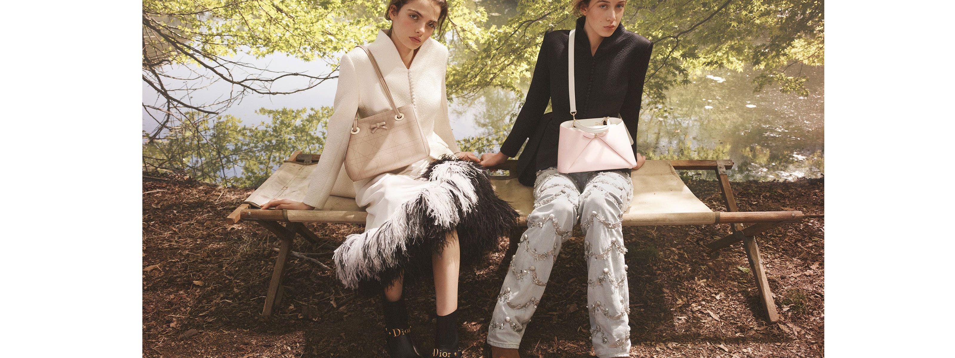 Two women seated outdoors on a wooden bench, wearing Dior tailoring and accessories in a wooded, waterside setting