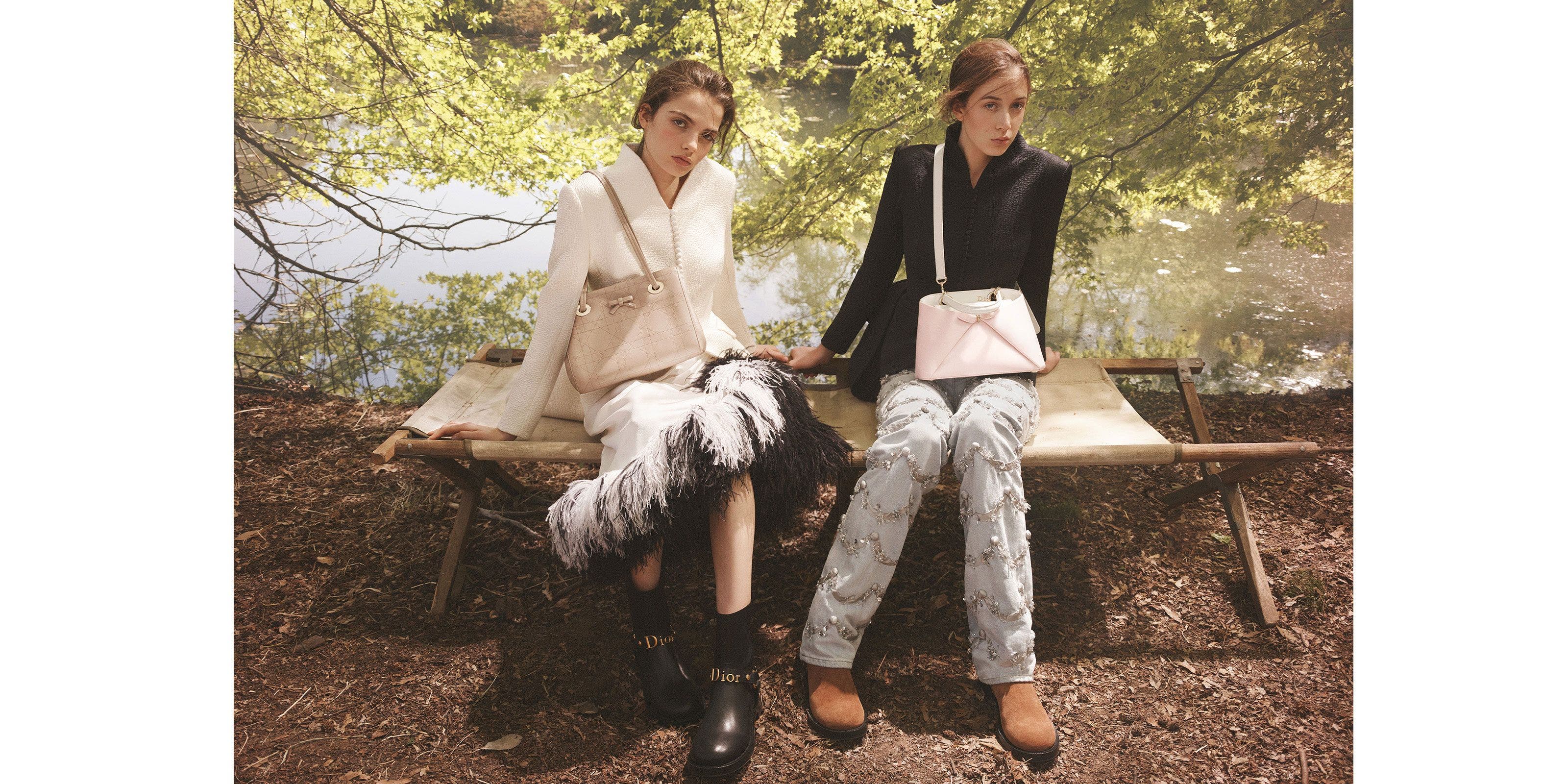 Two women seated outdoors on a wooden bench, wearing Dior tailoring and accessories in a wooded, waterside setting