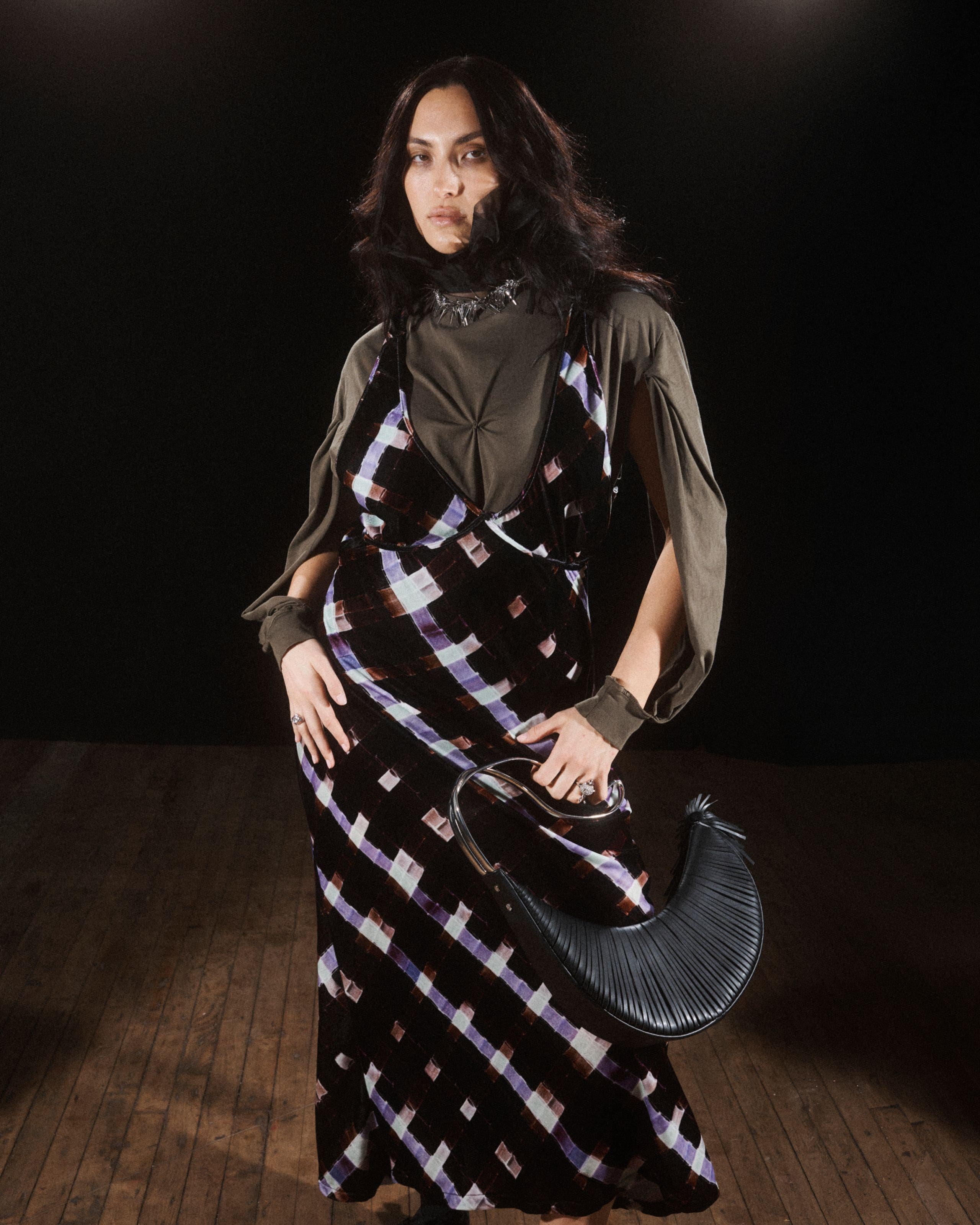 A model poses against a dark background, wearing a black slip-style maxi dress with a purple and white tie-dye or marbled pattern, layered over an olive-green long-sleeve top. She carries a black pleated clutch and stands on a wood floor.