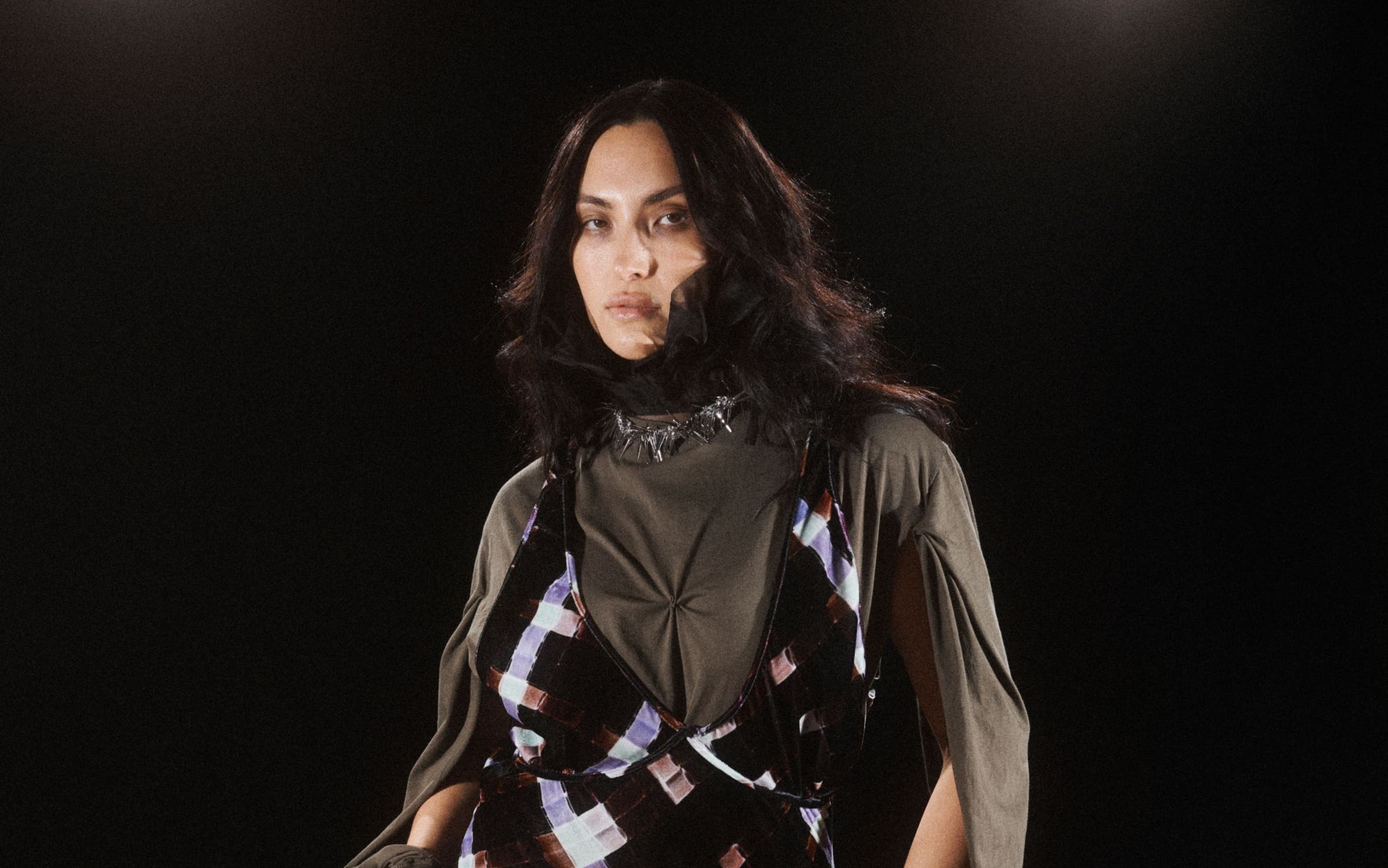 A model poses against a dark background, wearing a black slip-style maxi dress with a purple and white tie-dye or marbled pattern, layered over an olive-green long-sleeve top. She carries a black pleated clutch and stands on a wood floor.
