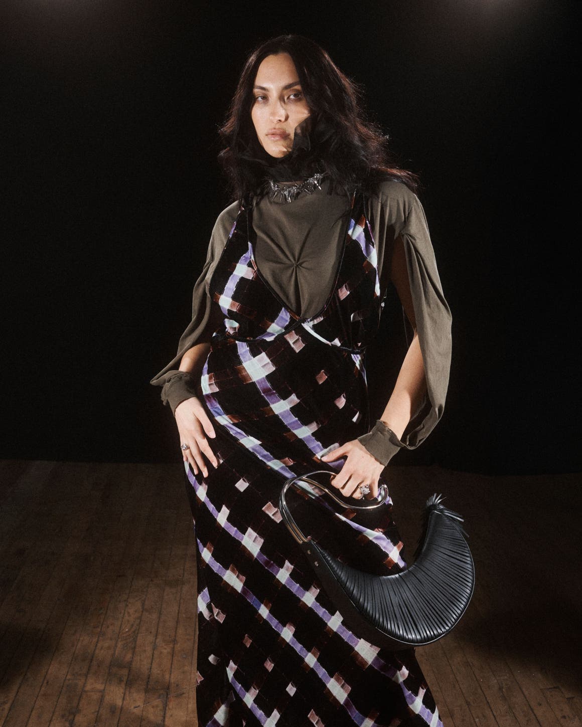 A model poses against a dark background, wearing a black slip-style maxi dress with a purple and white tie-dye or marbled pattern, layered over an olive-green long-sleeve top. She carries a black pleated clutch and stands on a wood floor.