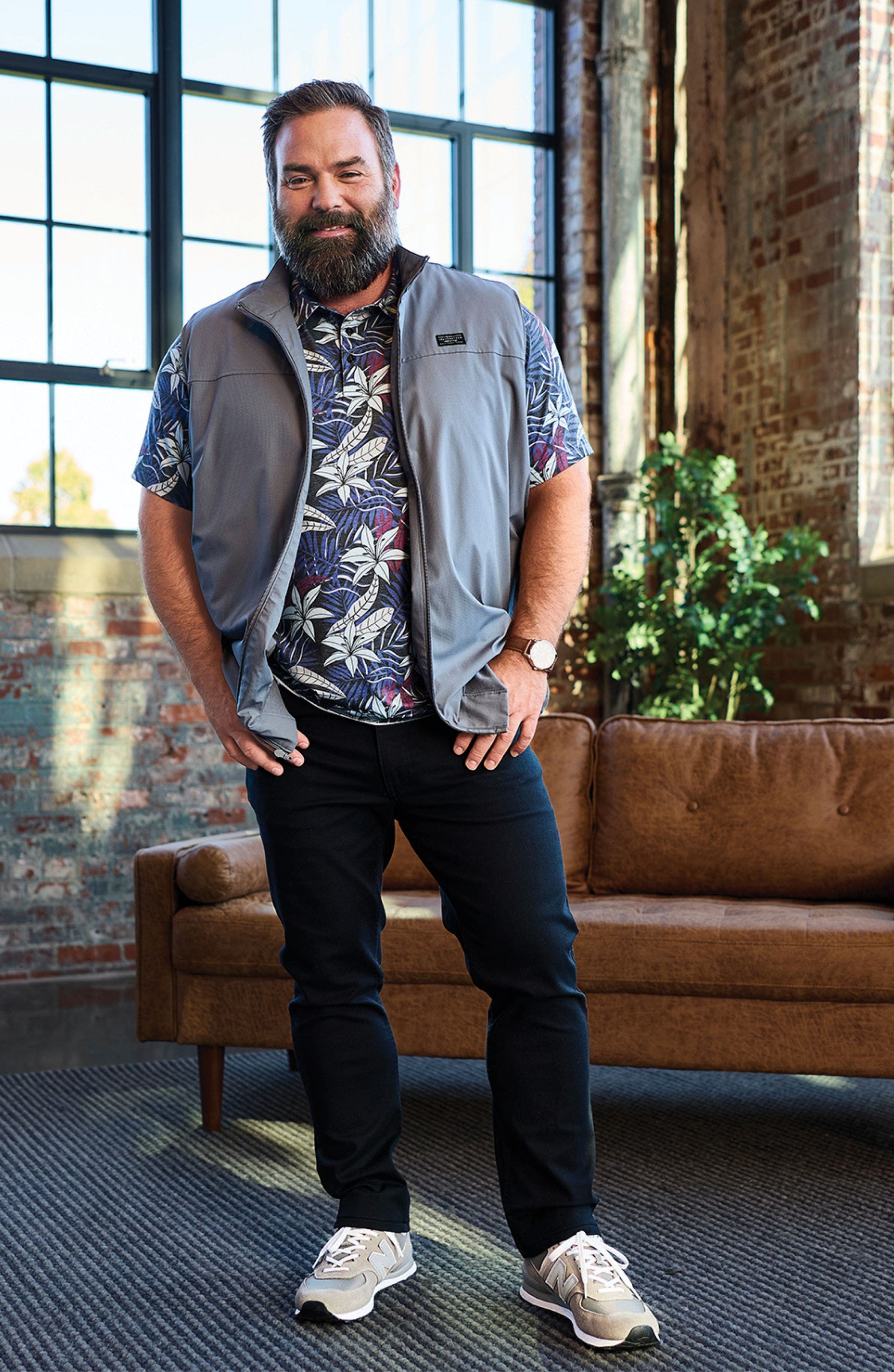A man wearing a light vest over a patterned short-sleeve shirt and dark pants.