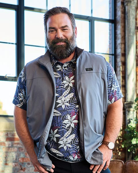 A man wearing a light vest over a patterned short-sleeve shirt and dark pants.