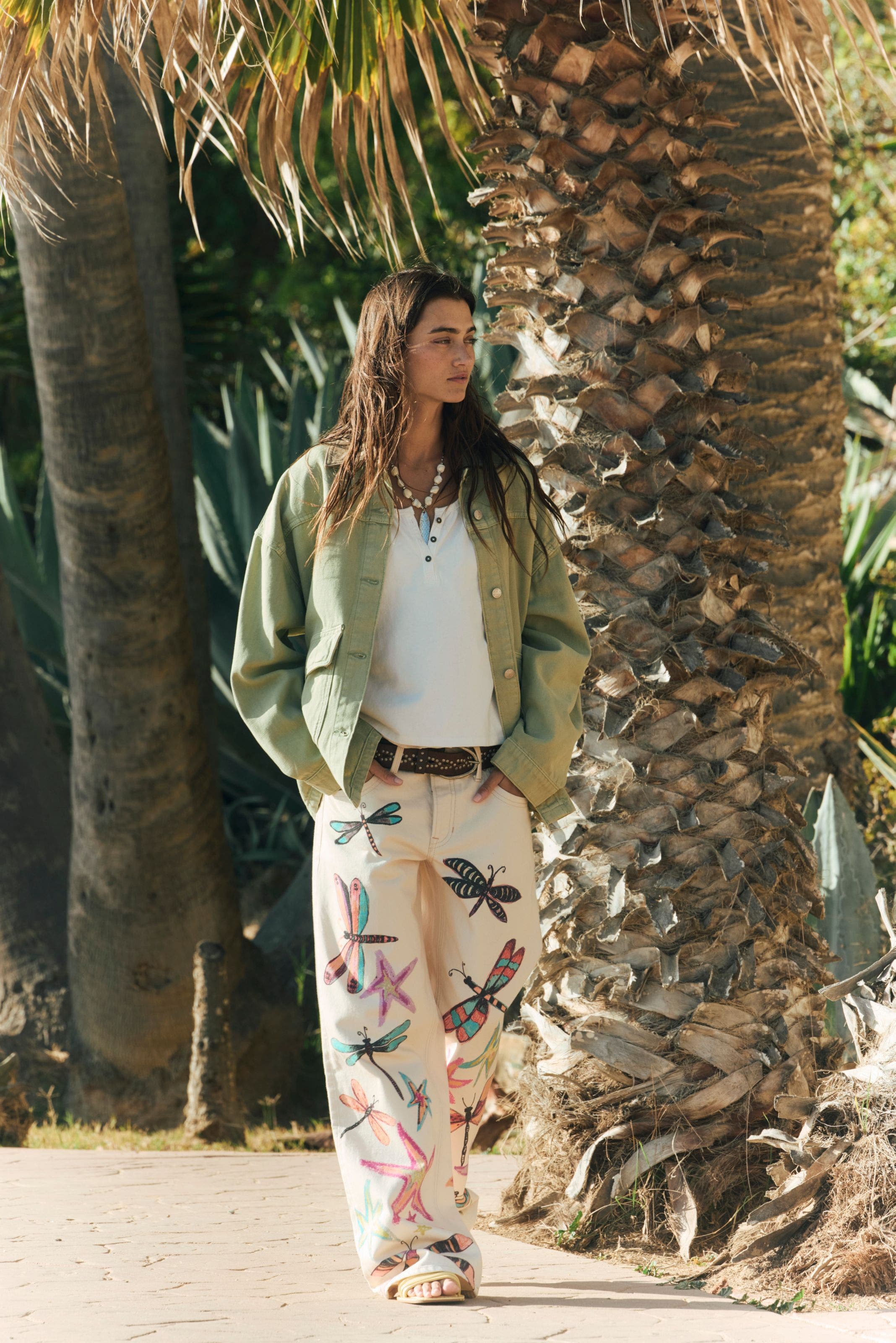 A woman wearing white jeans decorated with dragonflies and stars, paired with a white top and sage green jacket.