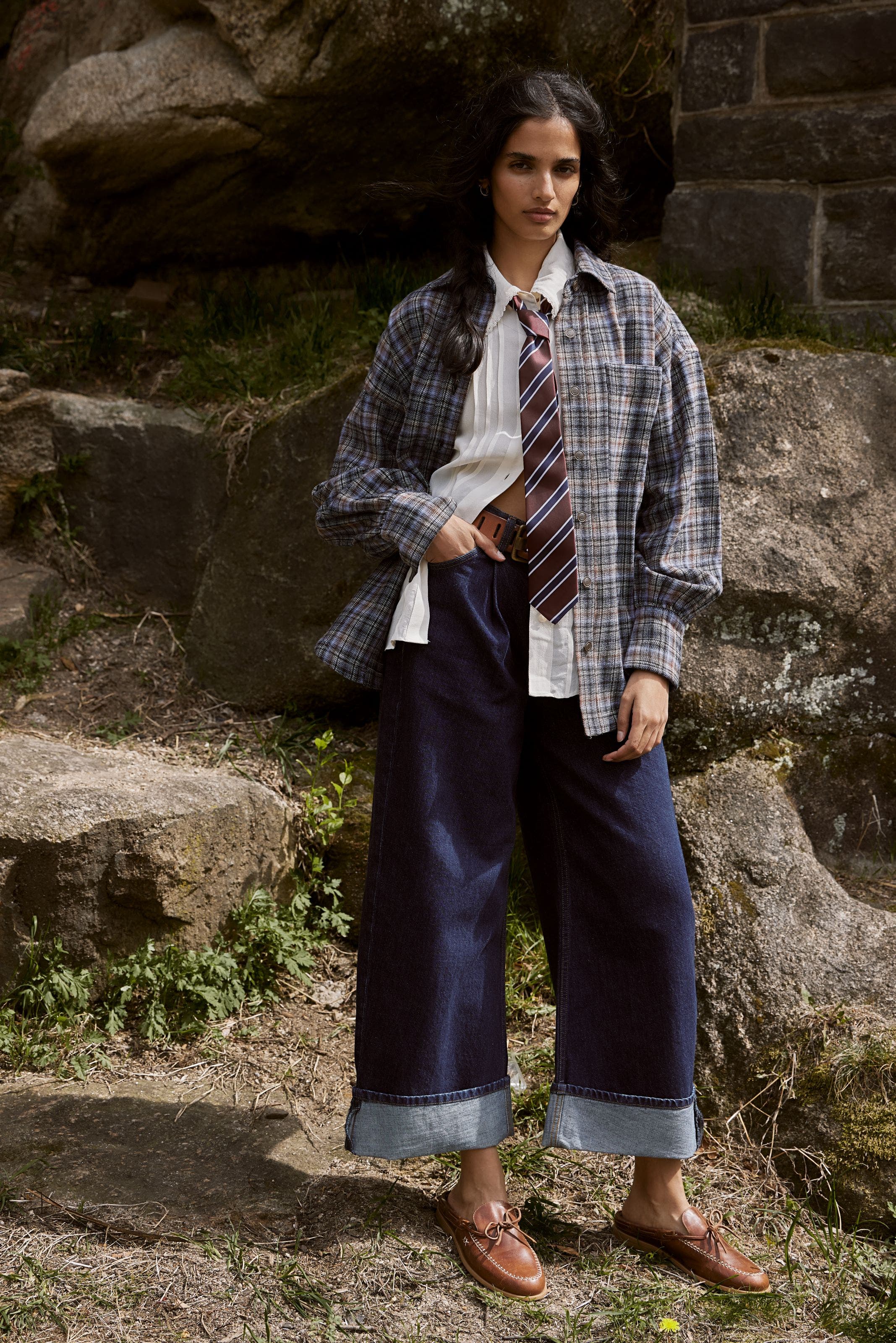 A woman wearing a plaid shirt, button up, tie and jeans.