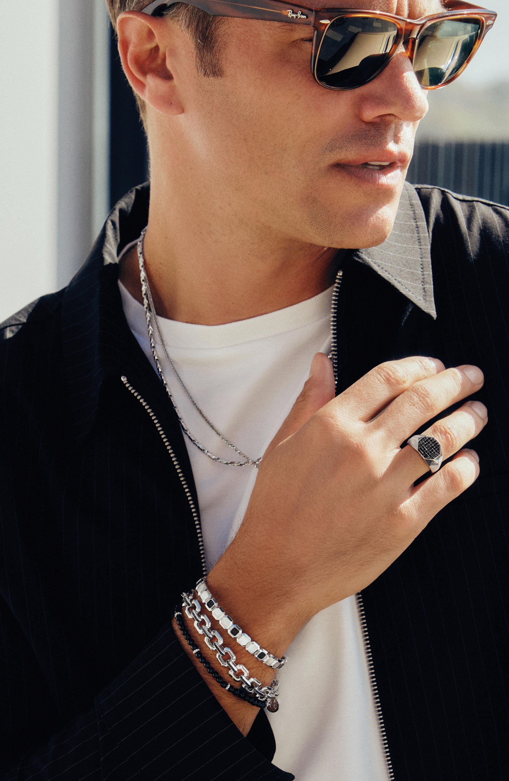 A man wearing multiple silver and beaded bracelets, chain necklaces and a ring.