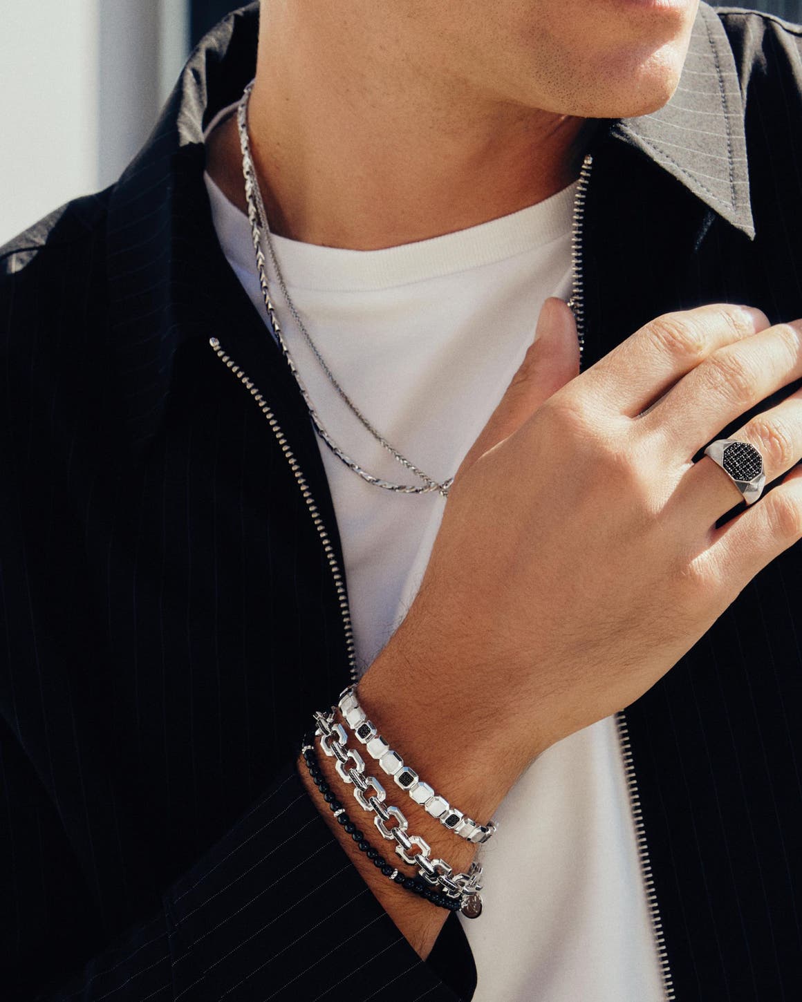 A man wearing multiple silver and beaded bracelets, chain necklaces and a ring.
