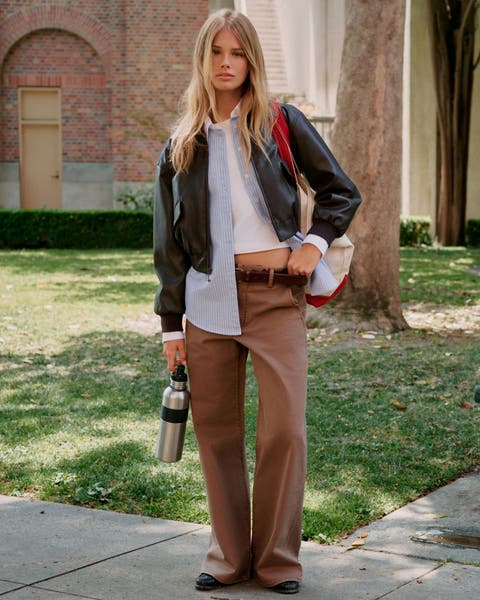 A woman wearing brown jeans, a white t-shirt, a blue Oxford shirt, ad a leather bomber.