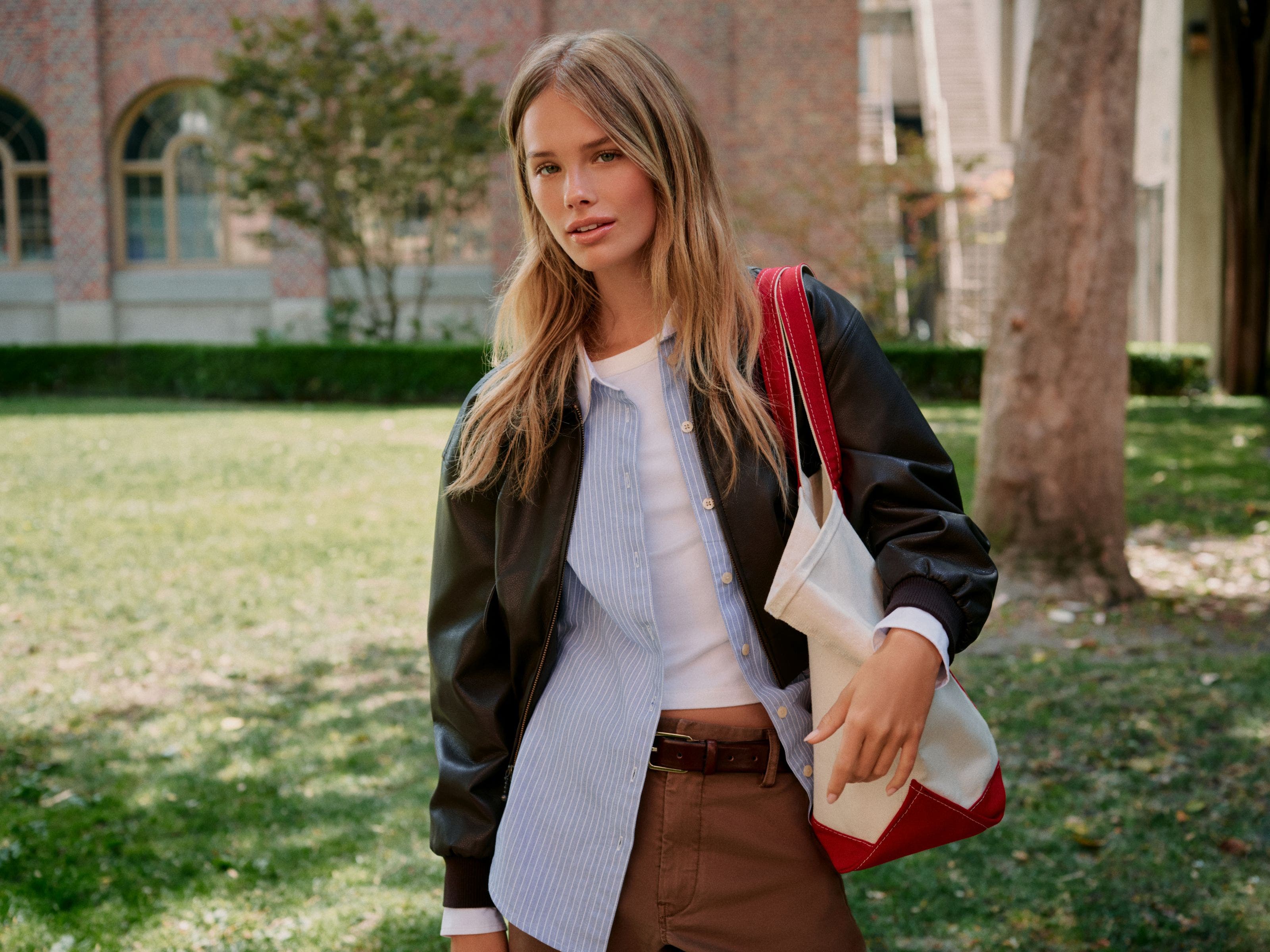 A woman wearing a white t-shirt, a blue button-up, brown slacks, and a leather bomber.