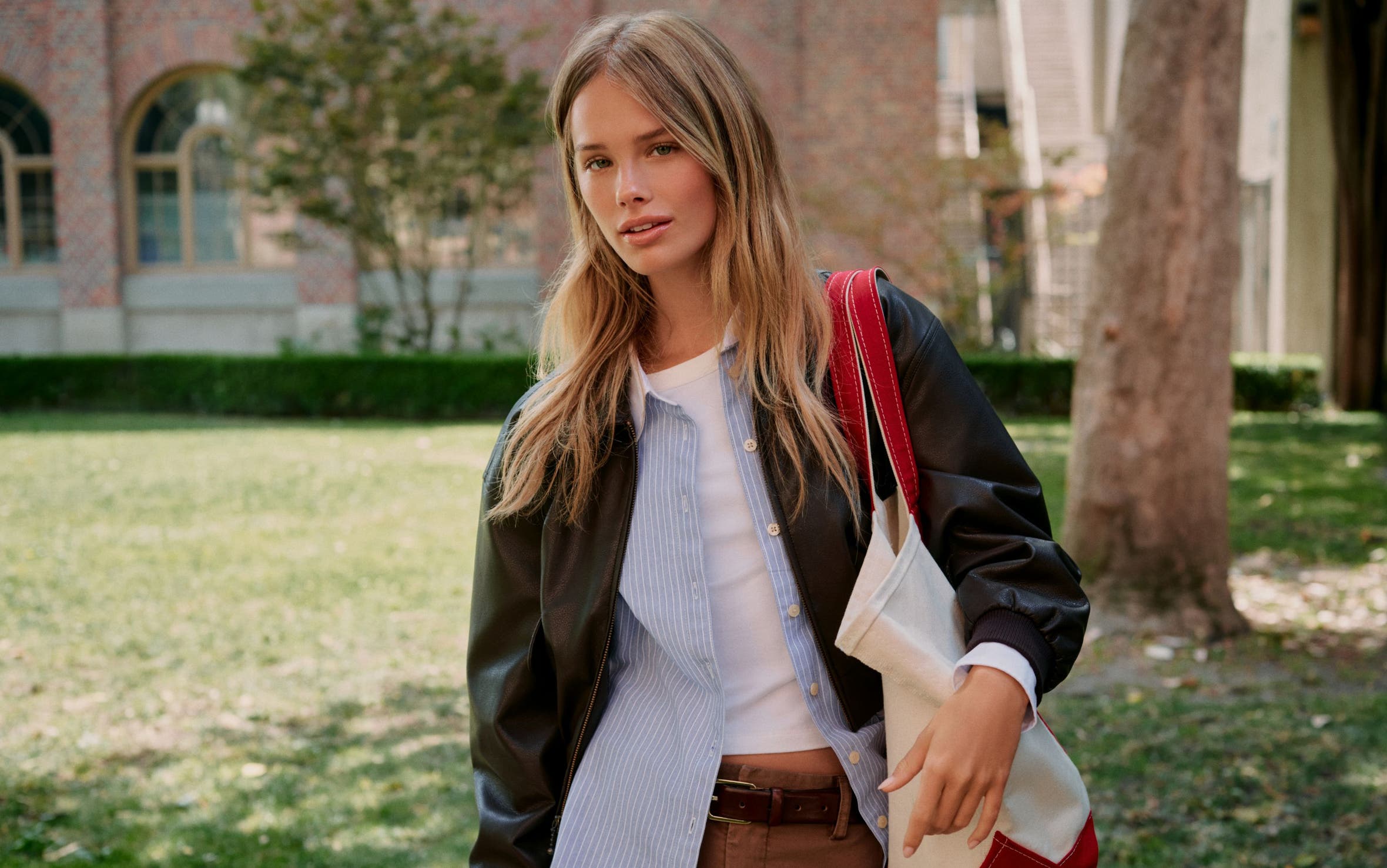 A woman wearing a white t-shirt, a blue button-up, brown slacks, and a leather bomber.