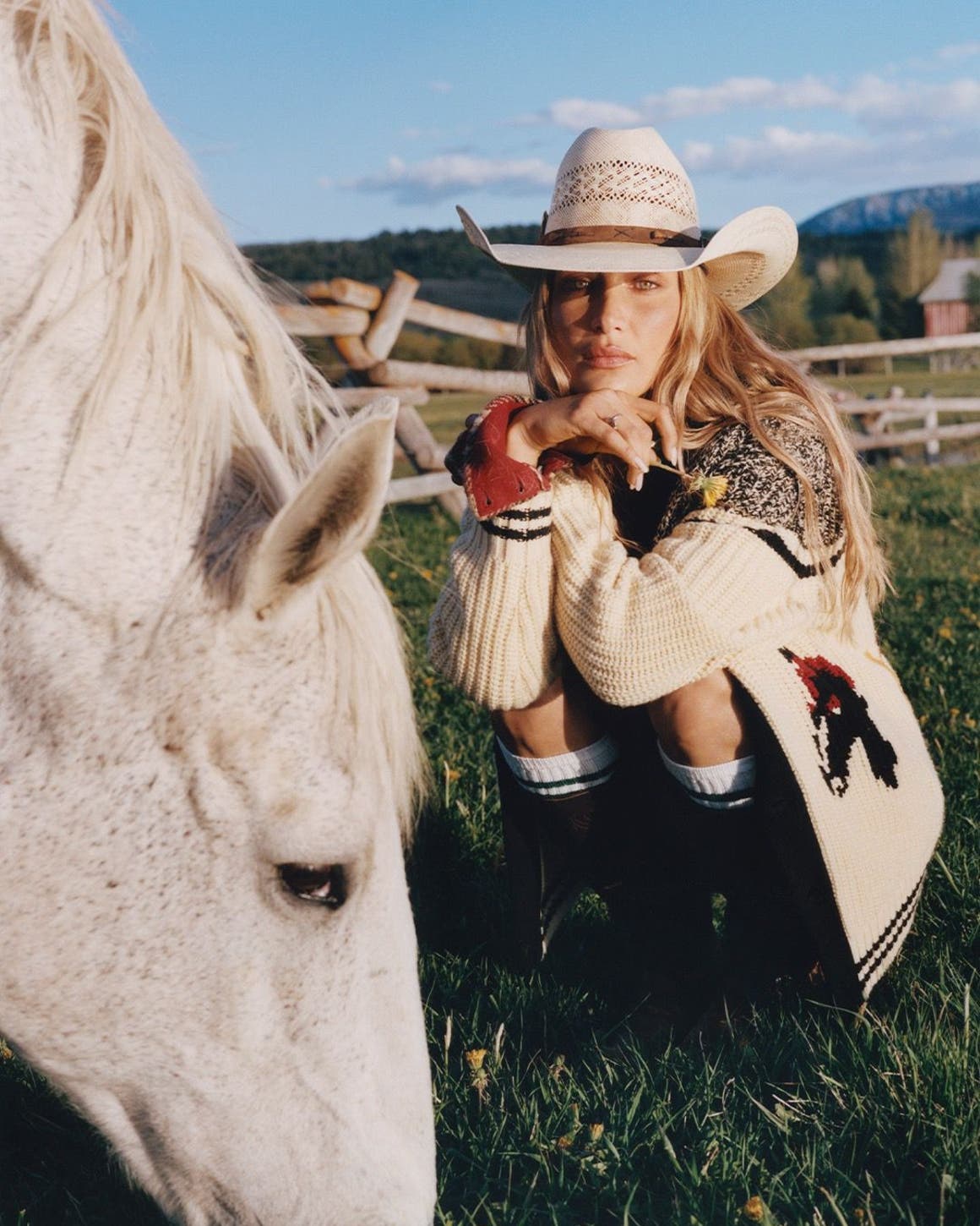 Bella Hadid wearing a chunky cardigan embroidered with horses and a cowgirl hat, sitting in a field with a horse.