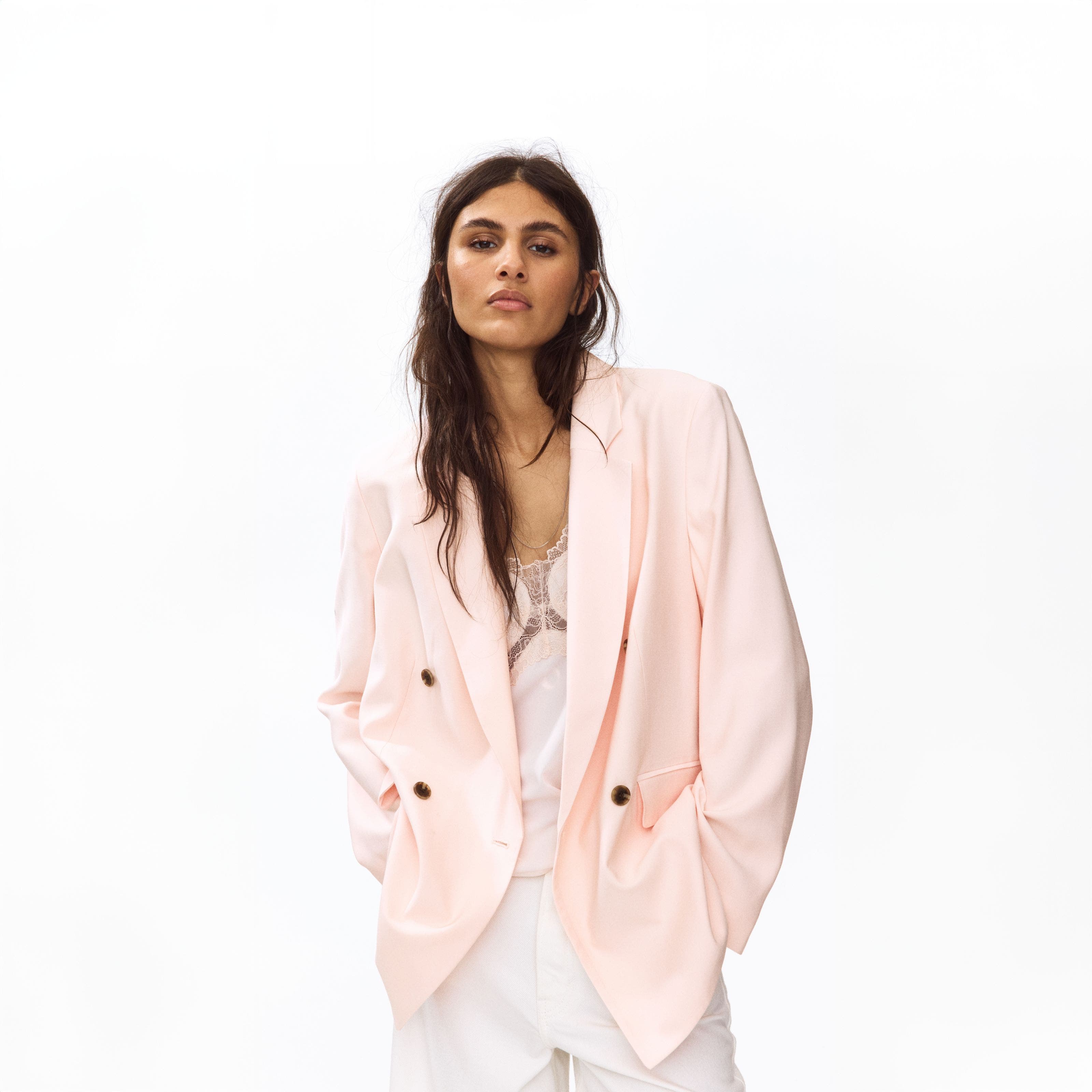 A woman wearing a light pink blazer over a white lace-trim camisole with white pants.