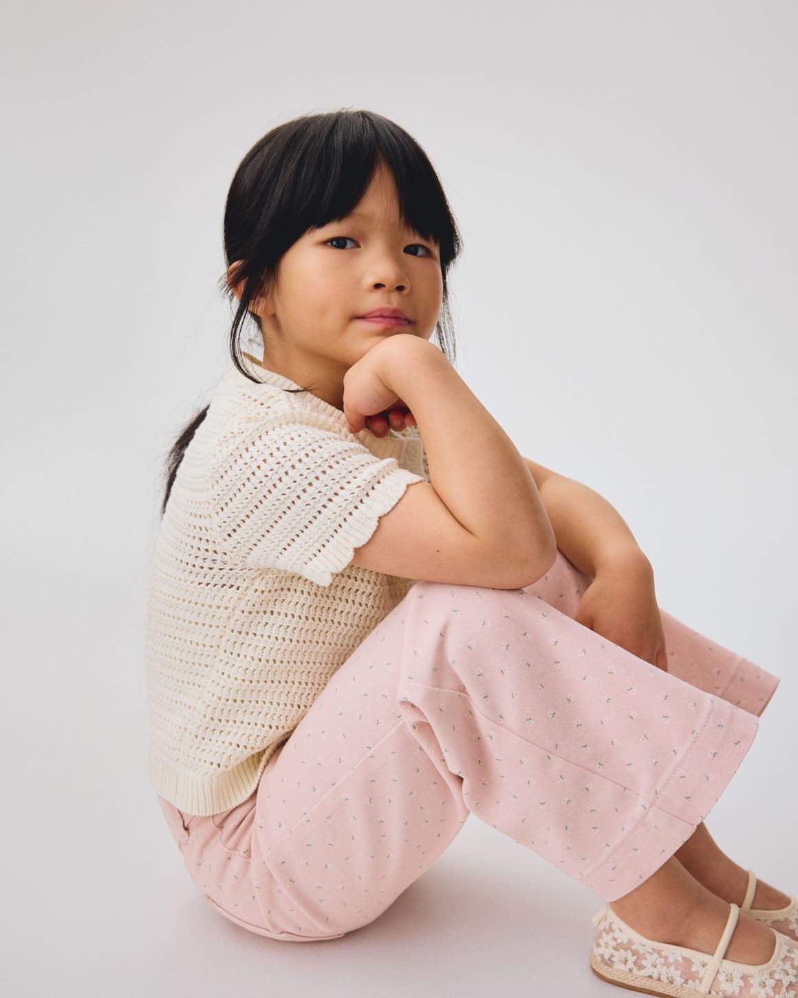 A girl wearing a cream scalloped cardigan and pink pants from H&M.
