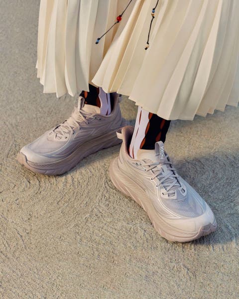 Model wearing lavender Hoka shoes with a white pleated skirt.