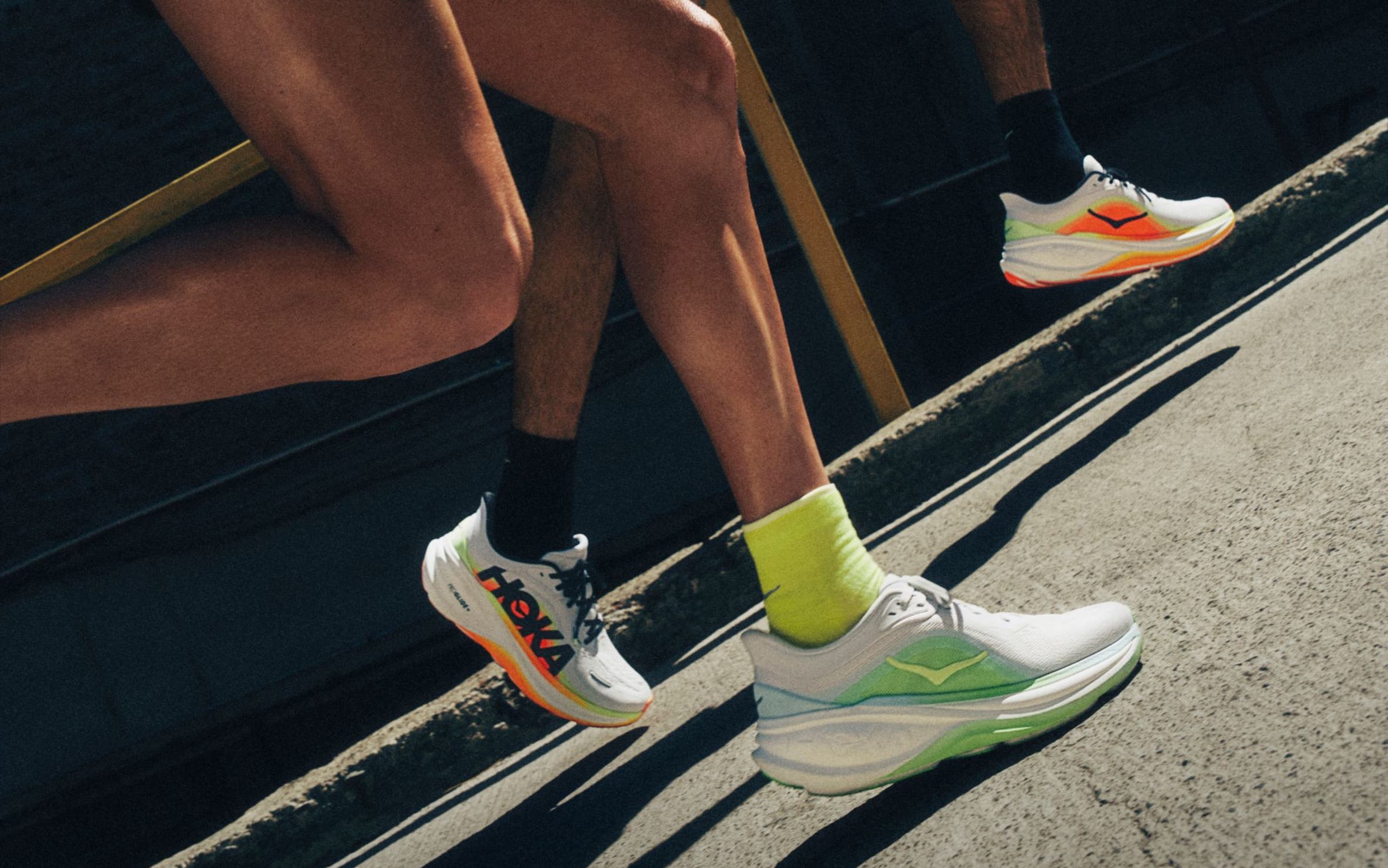 Runners on city pavement wearing orange and green HOKA sneakers.