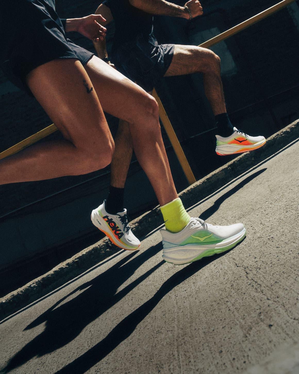 Runners on city pavement wearing orange and green HOKA sneakers.