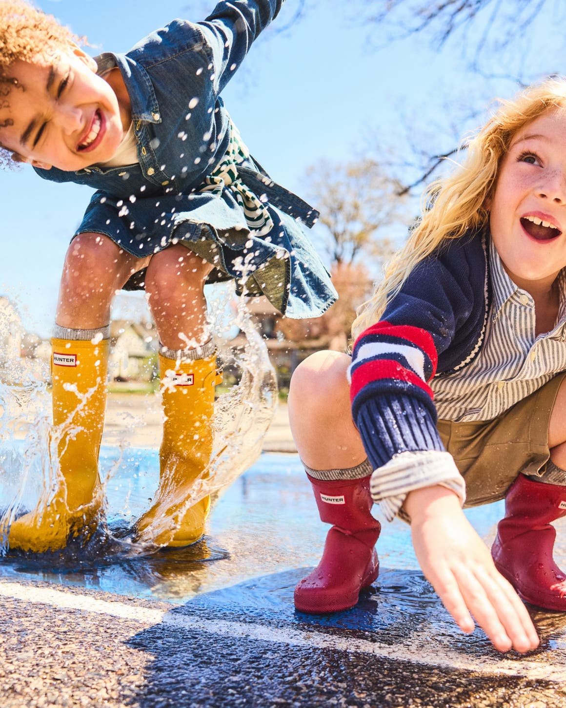 Two kids playing and splashing in a puddle while wearing colorful rain boots.