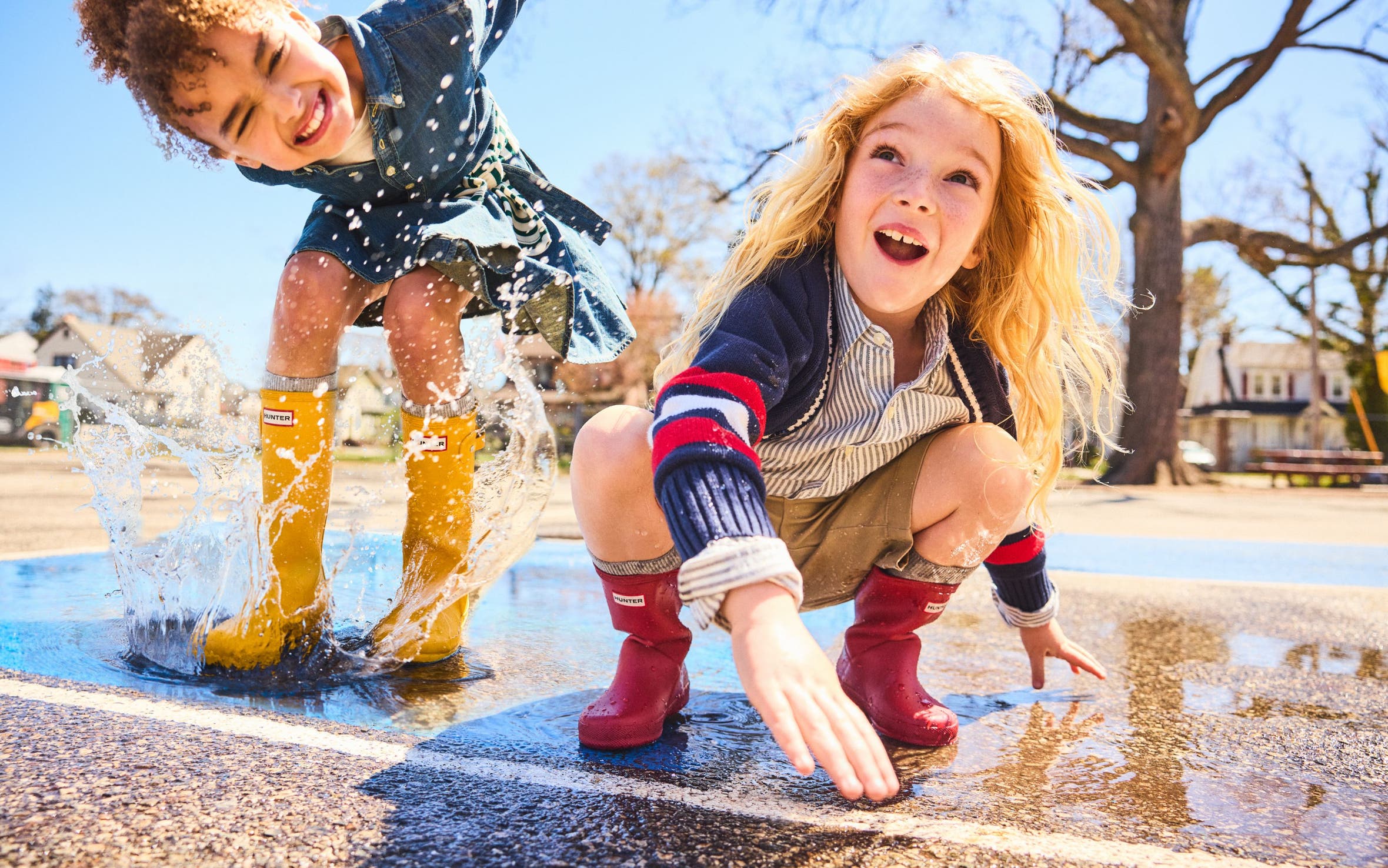 Two kids playing and splashing in a puddle while wearing colorful rain boots.