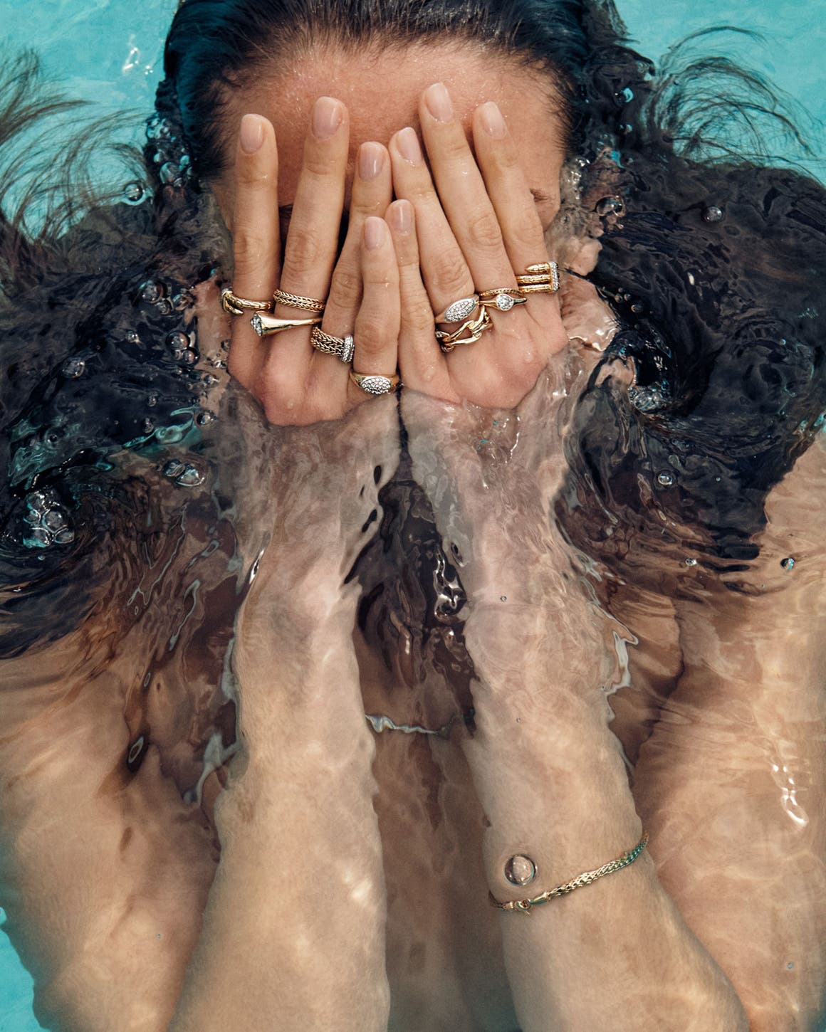 A woman swimming in a pool wearing stacks of John Hardy rings on each finger and a bracelet on one wrist.