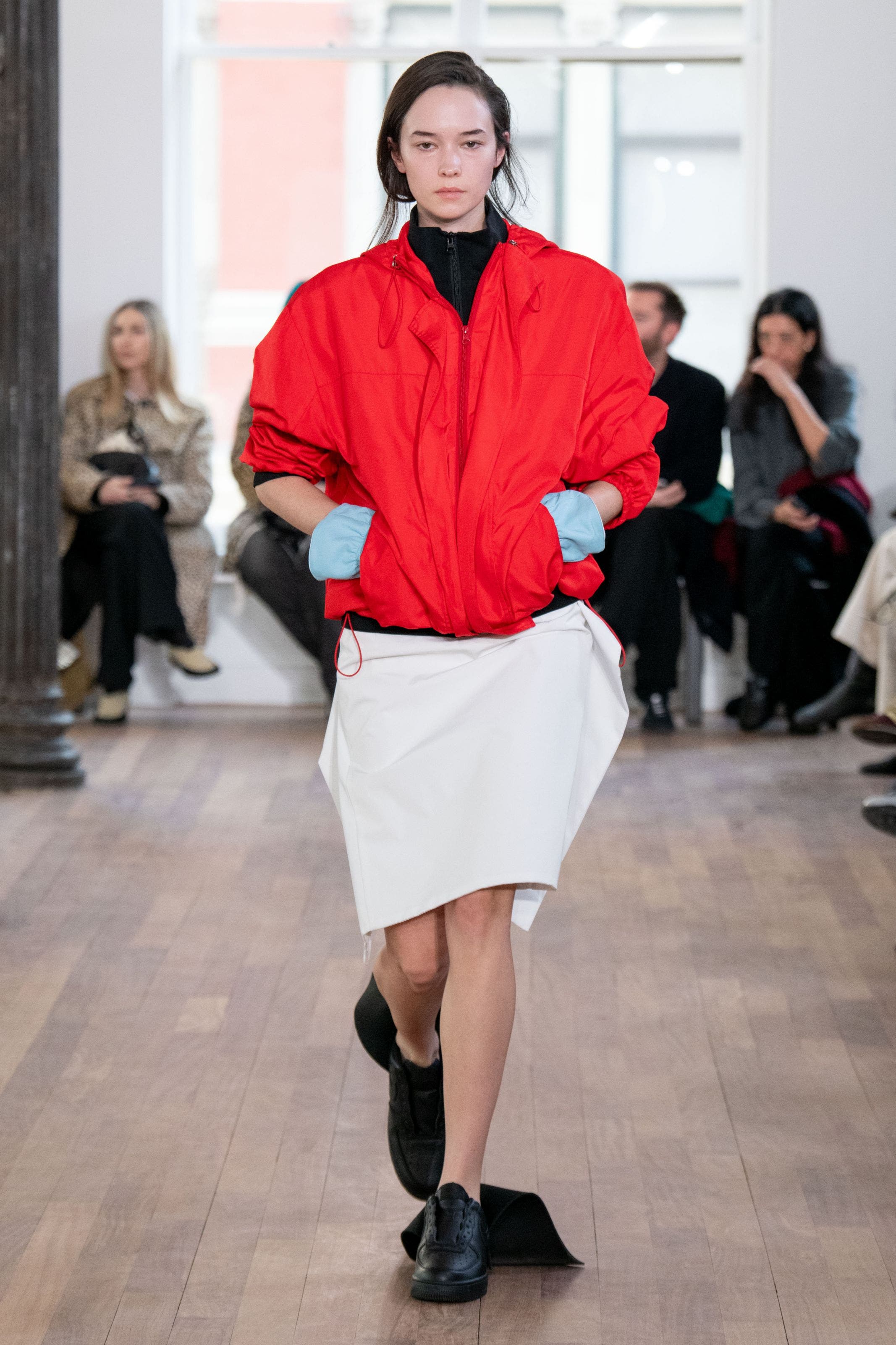 Model walks a runway wearing an oversized red jacket over a black turtleneck, paired with a cream midi skirt and black boots
