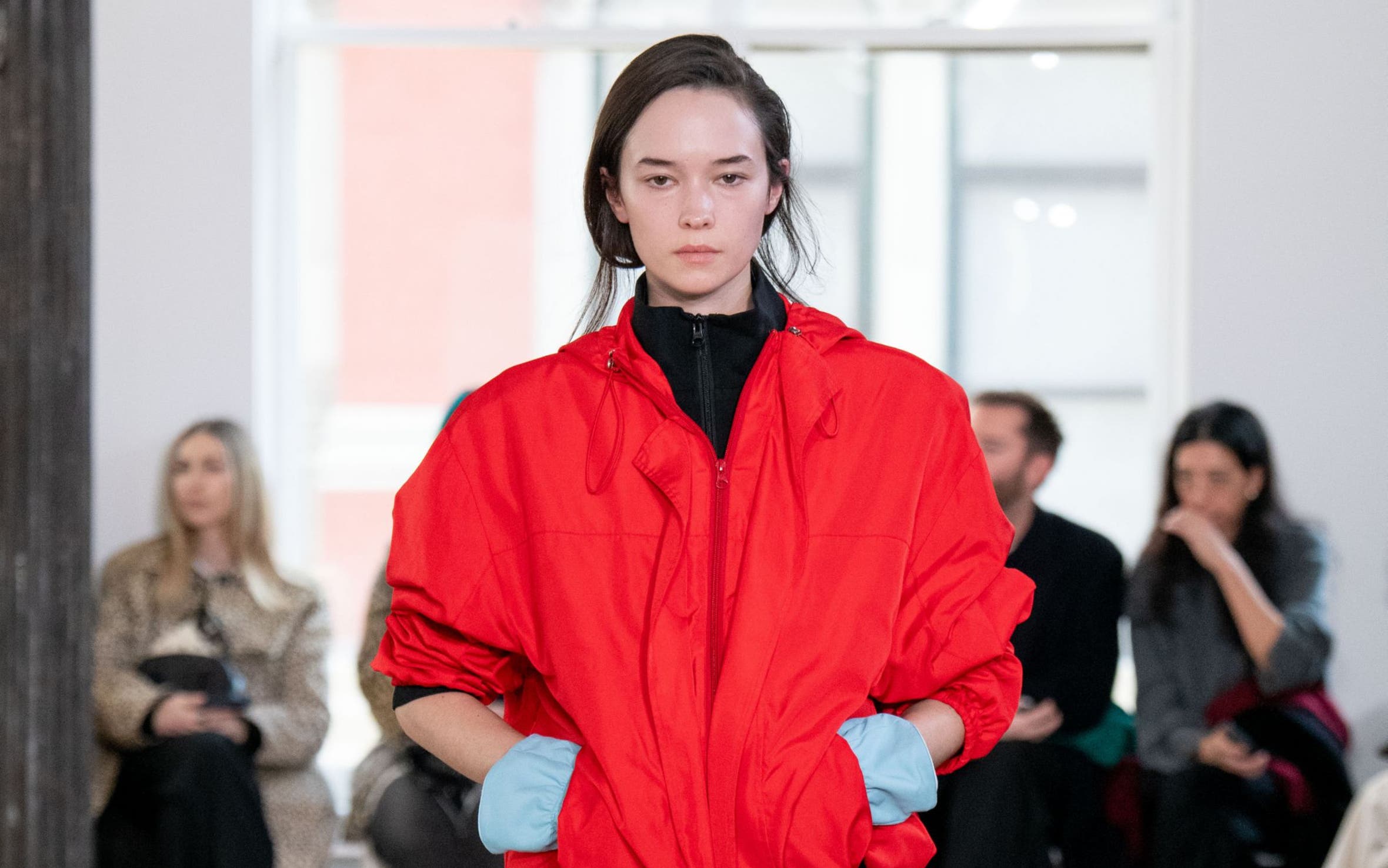 Model walks a runway wearing an oversized red jacket over a black turtleneck, paired with a cream midi skirt and black boots