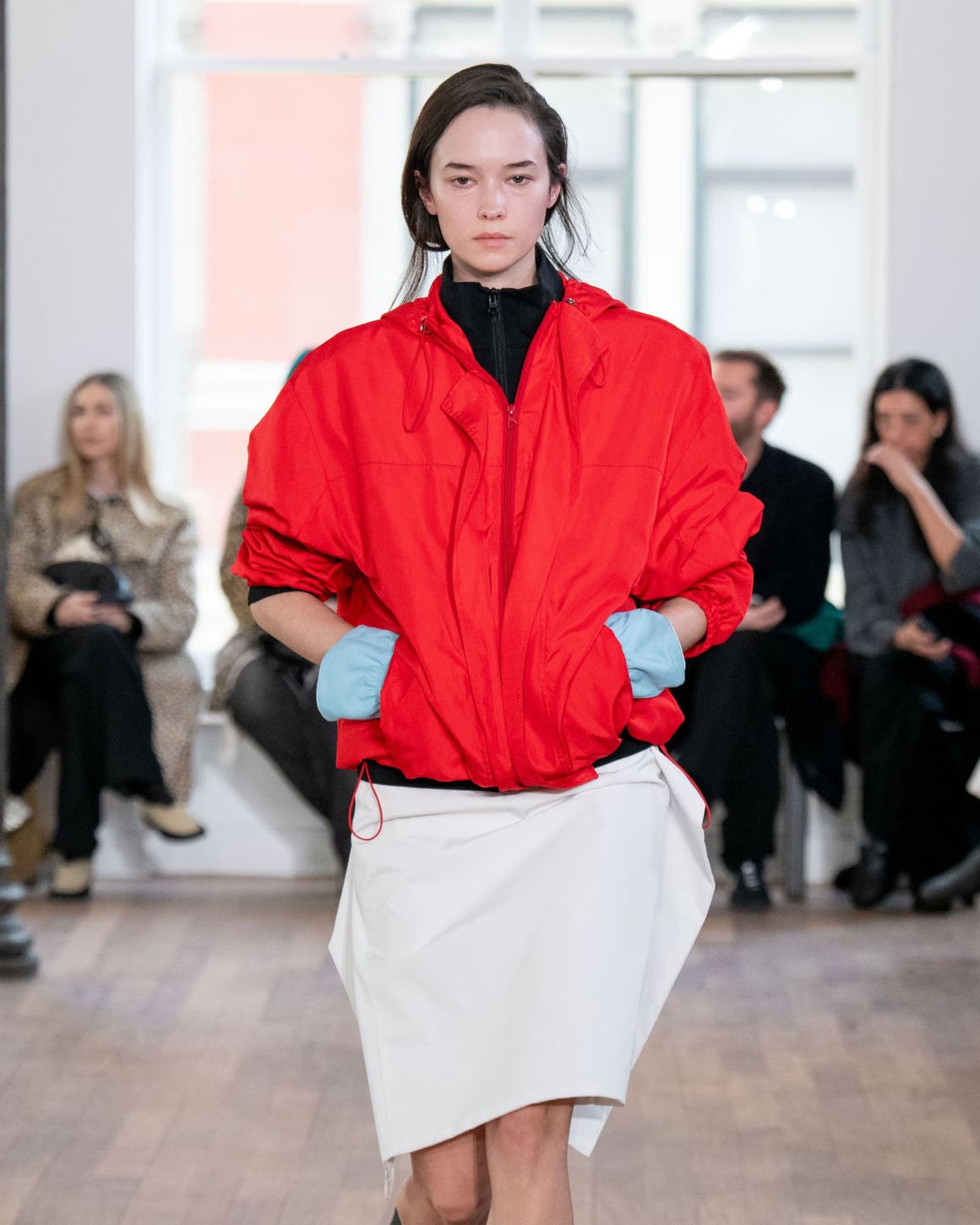 Model walks a runway wearing an oversized red jacket over a black turtleneck, paired with a cream midi skirt and black boots
