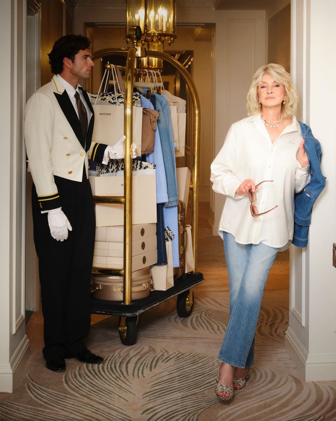Martha Stewart wearing a white button-up shirt, jeans and carrying a denim jacket next to a bellhop with a rack of boxes and clothes.