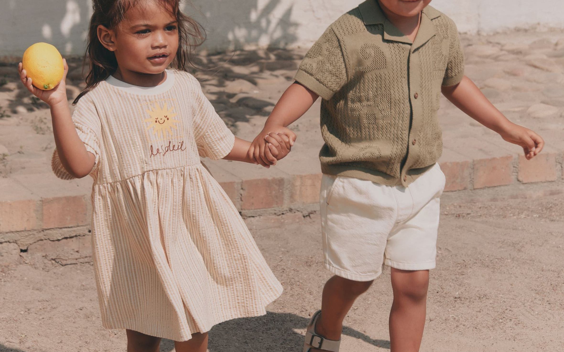 Two young children holding hands outdoors, dressed in neutral linen separates — a girl in a cream dress and a boy in a sage knit top with white shorts.