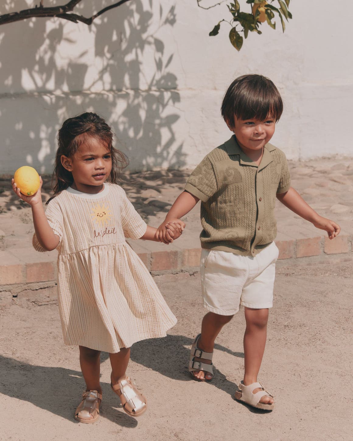 Two young children holding hands outdoors, dressed in neutral linen separates — a girl in a cream dress and a boy in a sage knit top with white shorts.