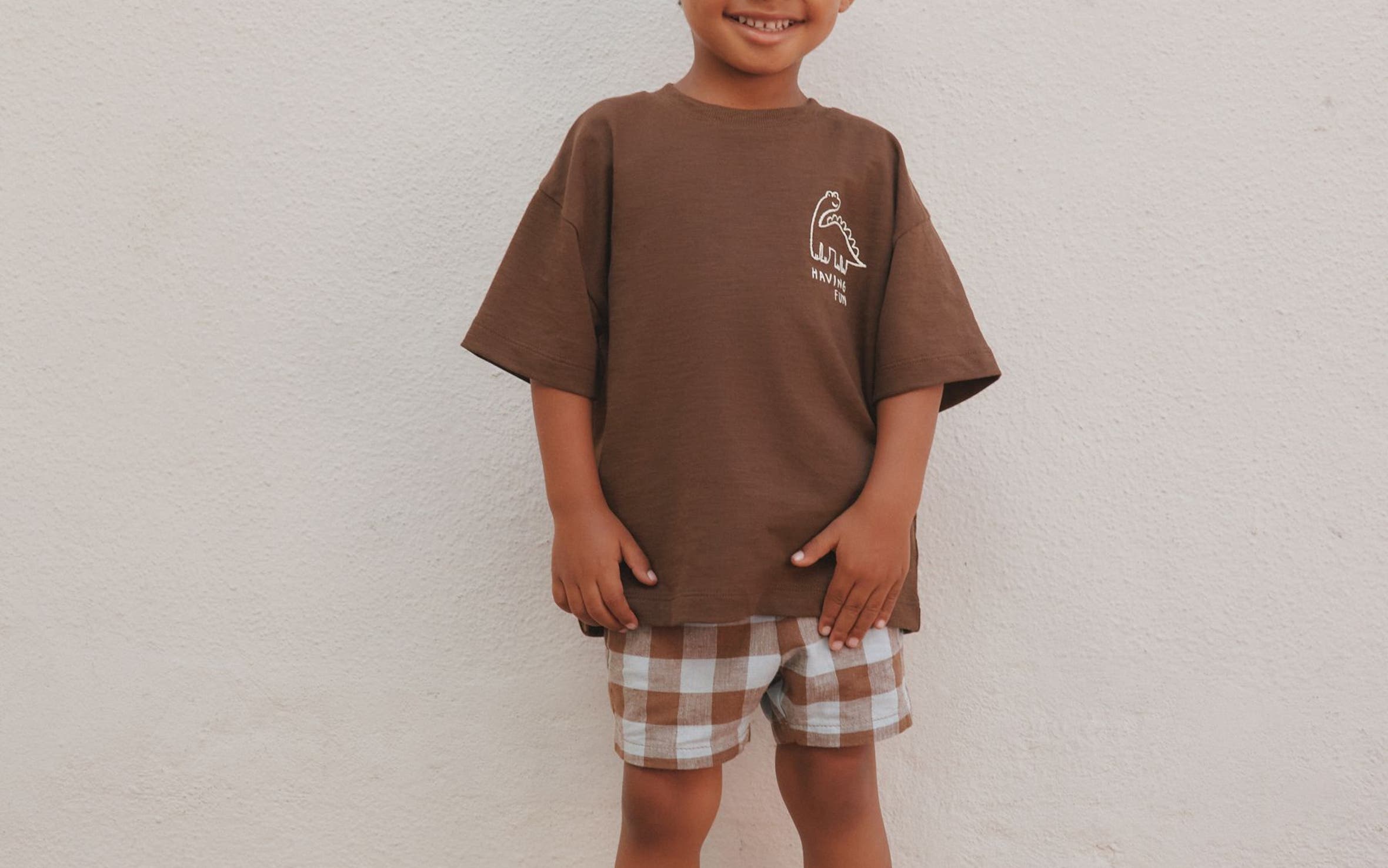 A young boy smiling in an oversized brown graphic T-shirt and brown-and-white plaid shorts, standing against a white wall.