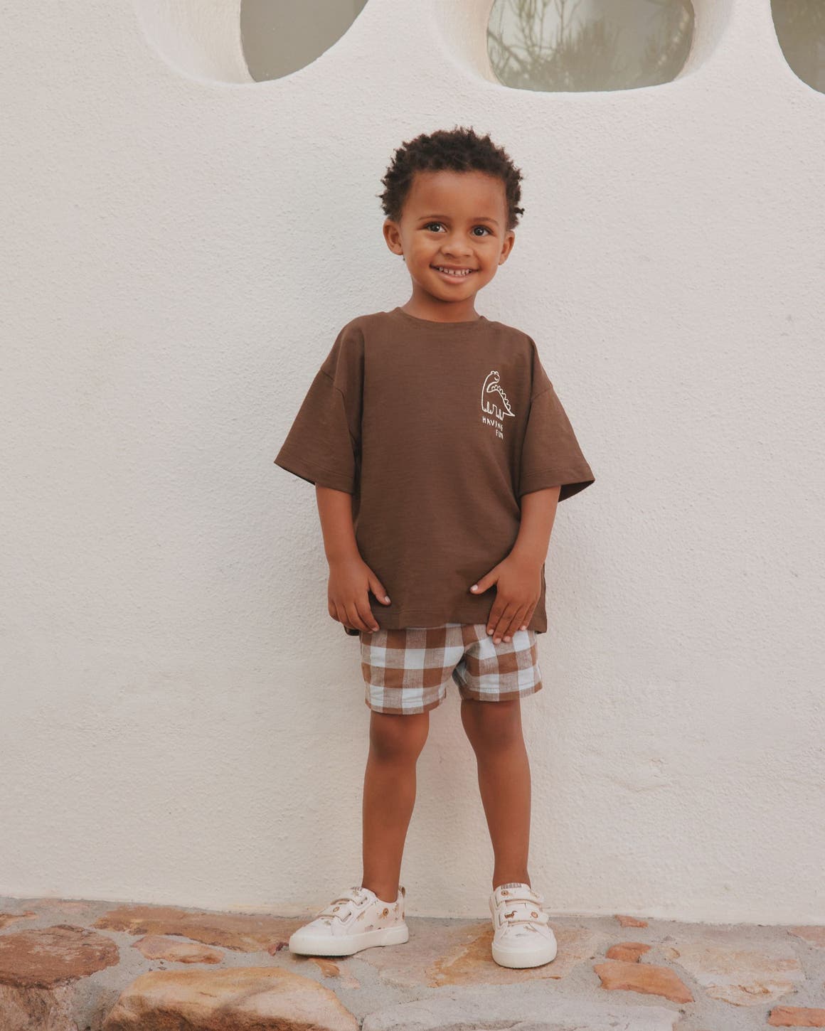 A young boy smiling in an oversized brown graphic T-shirt and brown-and-white plaid shorts, standing against a white wall.