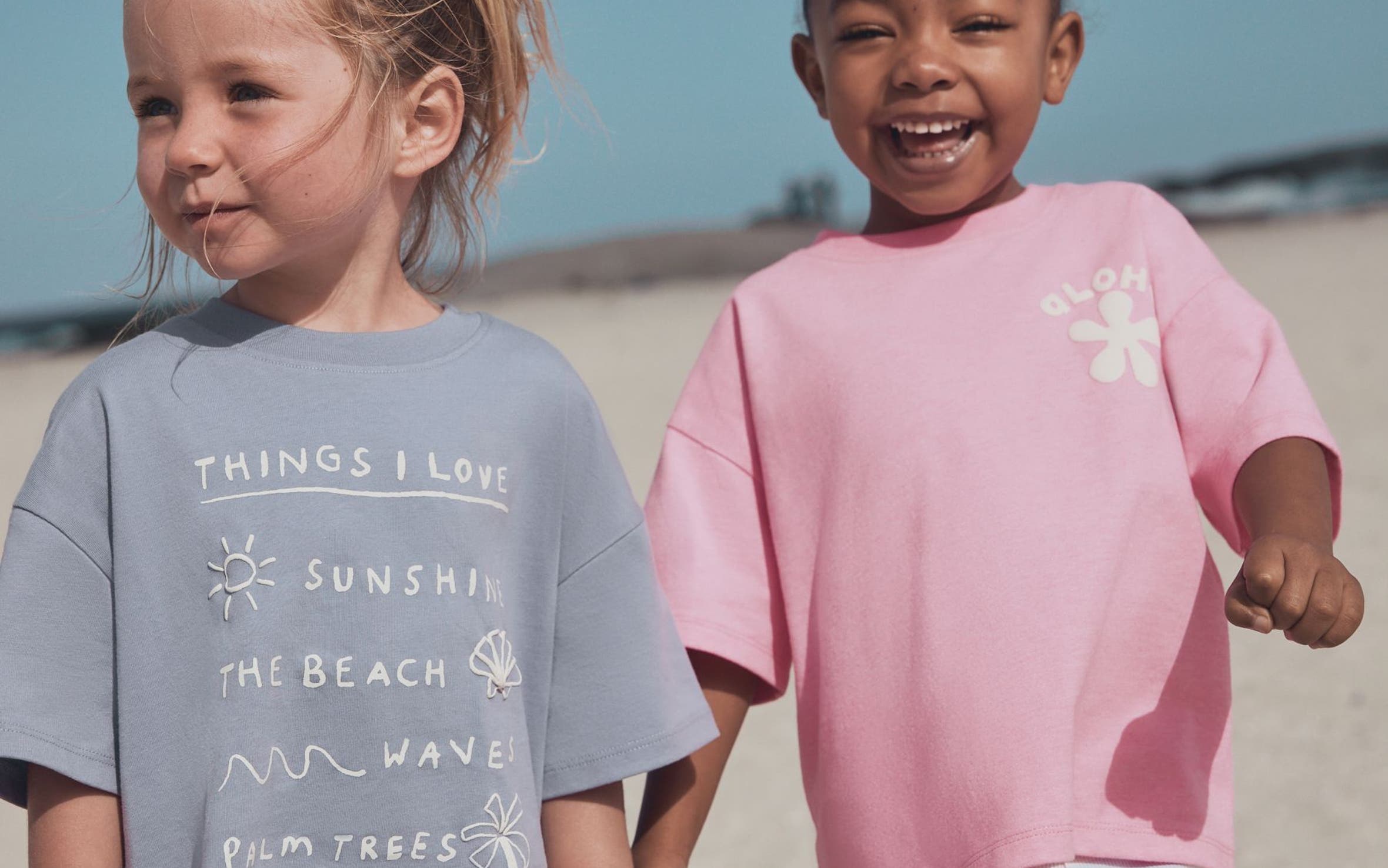 Two young girls holding hands on a beach, one in a blue graphic tee with floral shorts, the other in a pink tee with striped shorts.