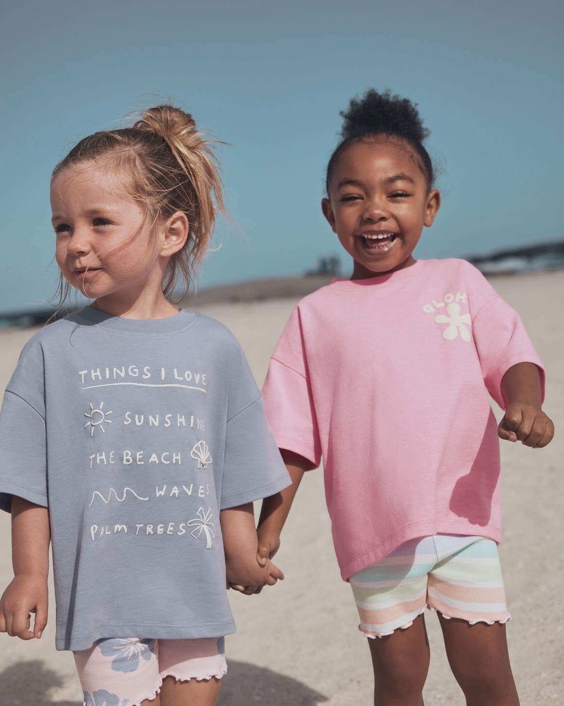 Two young girls holding hands on a beach, one in a blue graphic tee with floral shorts, the other in a pink tee with striped shorts.