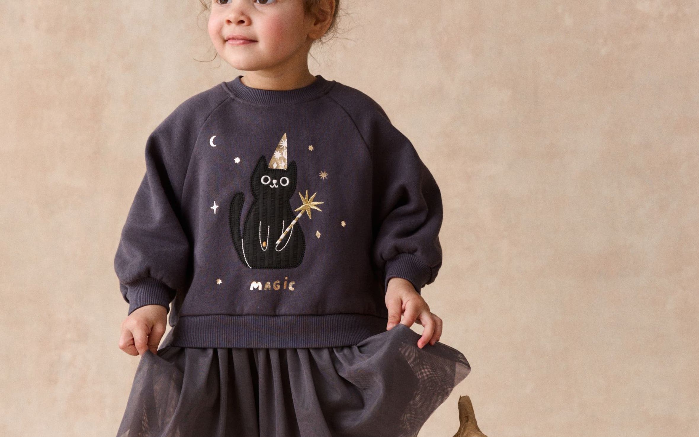 A girl in a sweatshirt with a black cat "Magic" graphic, a tulle skirt, boots and socks, standing next to a white pumpkin. 