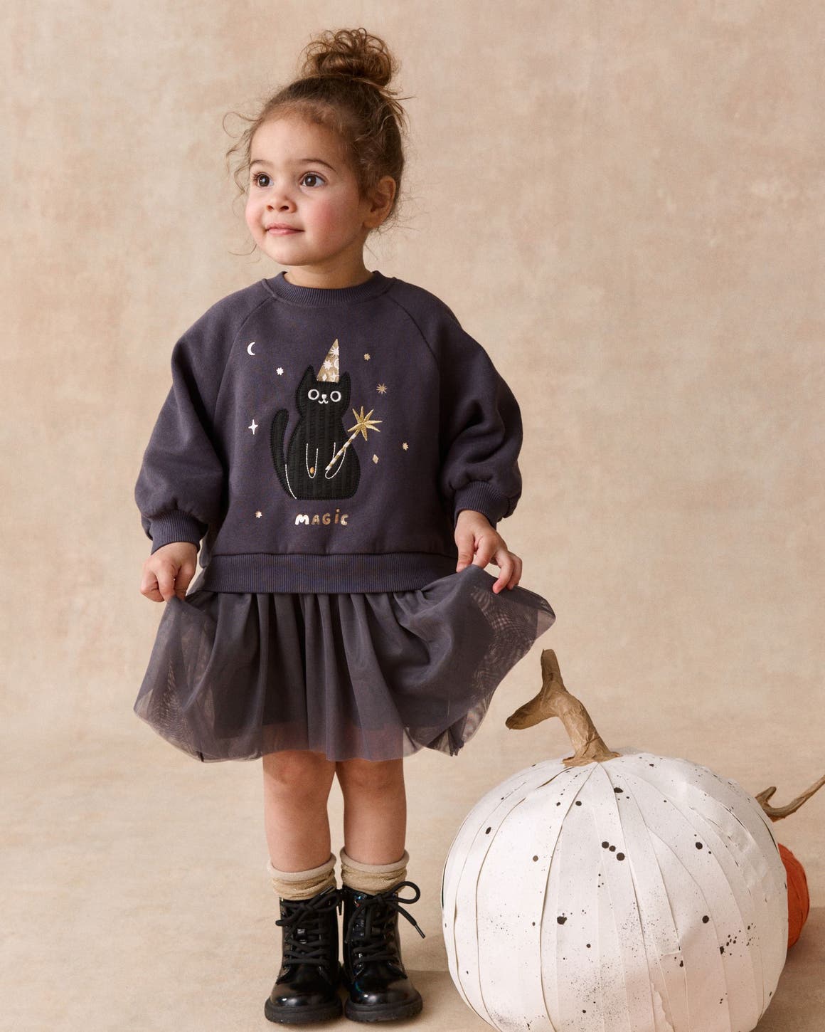 A girl in a sweatshirt with a black cat "Magic" graphic, a tulle skirt, boots and socks, standing next to a white pumpkin. 
