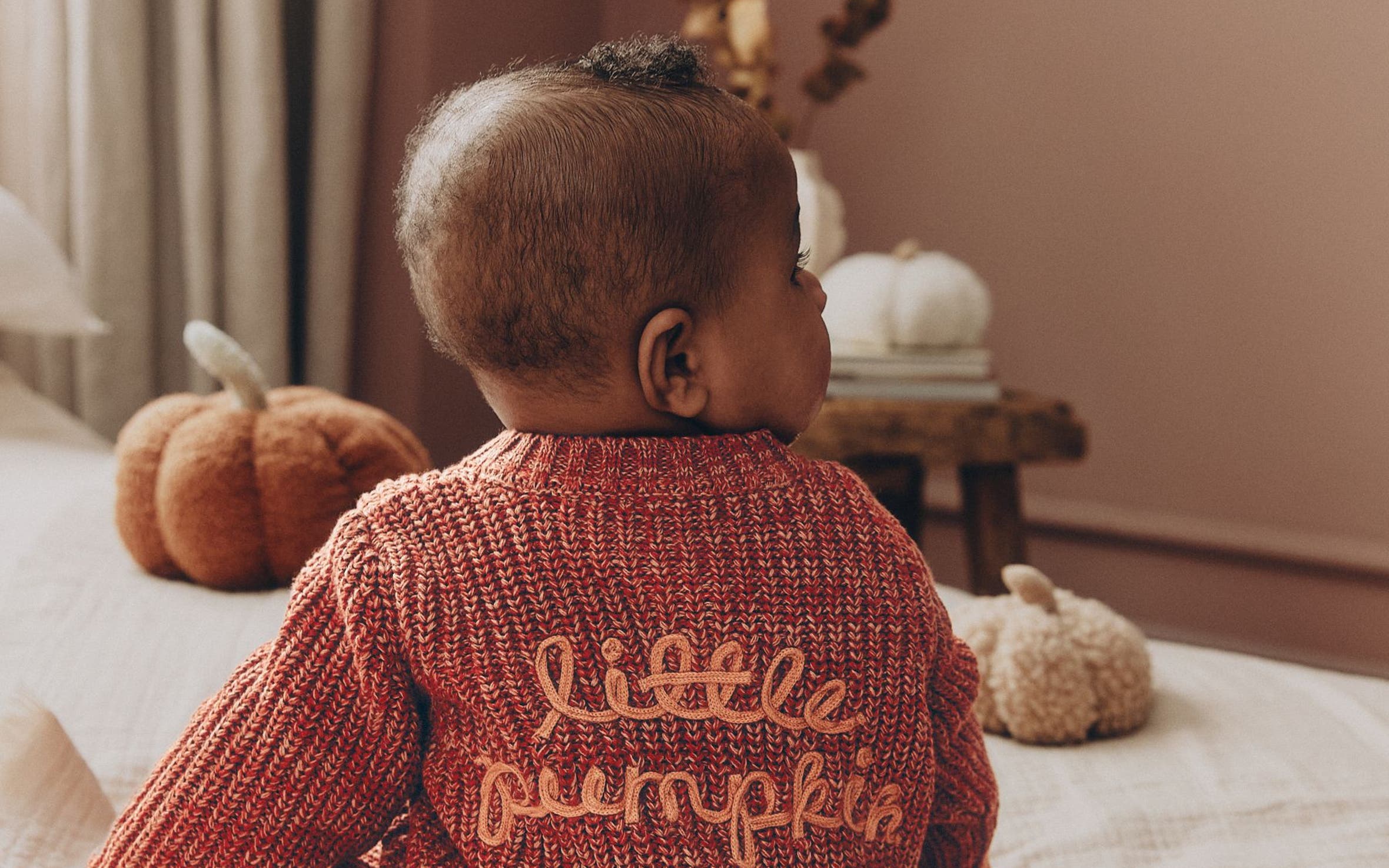 A baby in a rust-colored knit sweater embroidered "little pumpkin," sitting on a bed with plush pumpkins nearby.