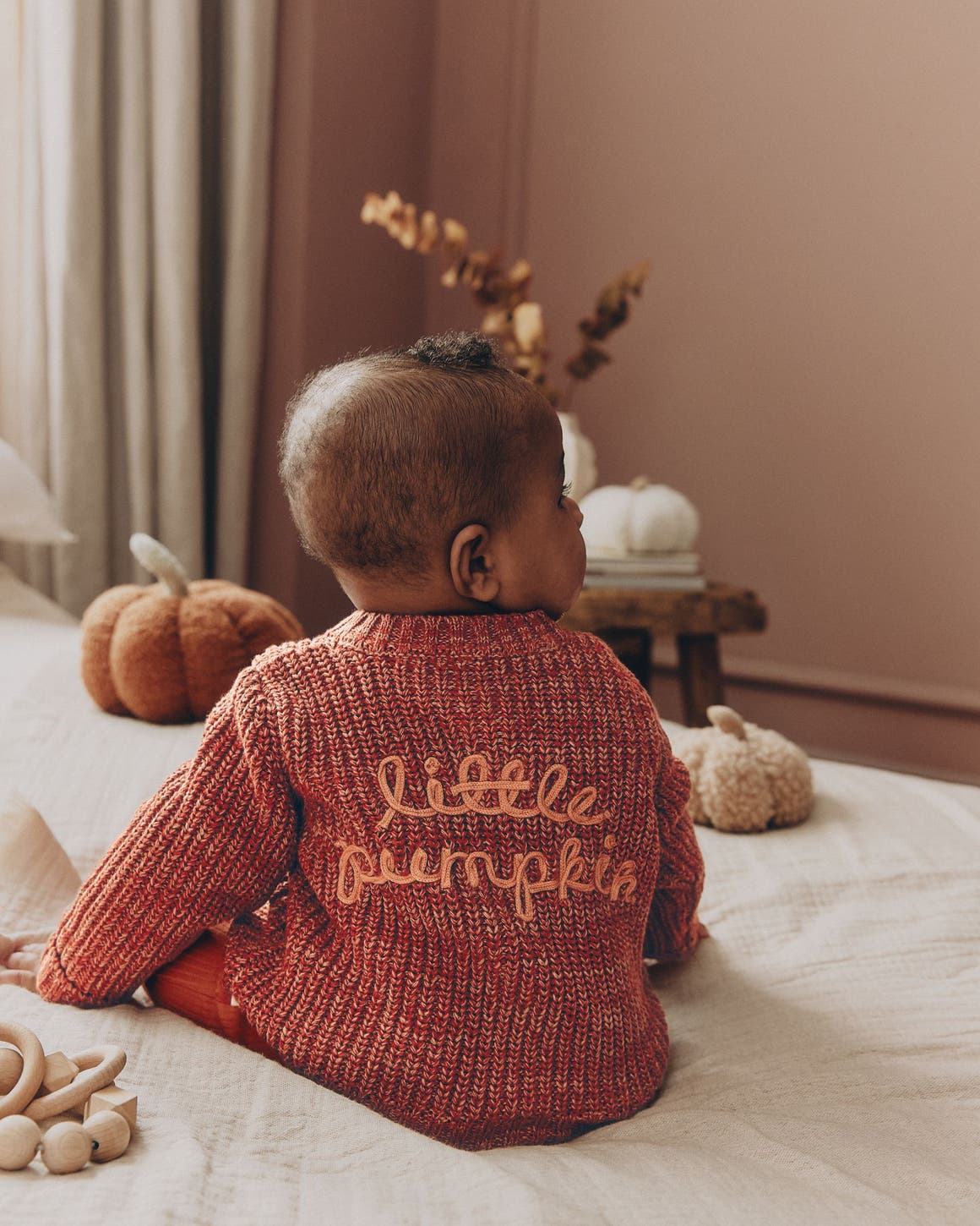 A baby in a rust-colored knit sweater embroidered "little pumpkin," sitting on a bed with plush pumpkins nearby.
