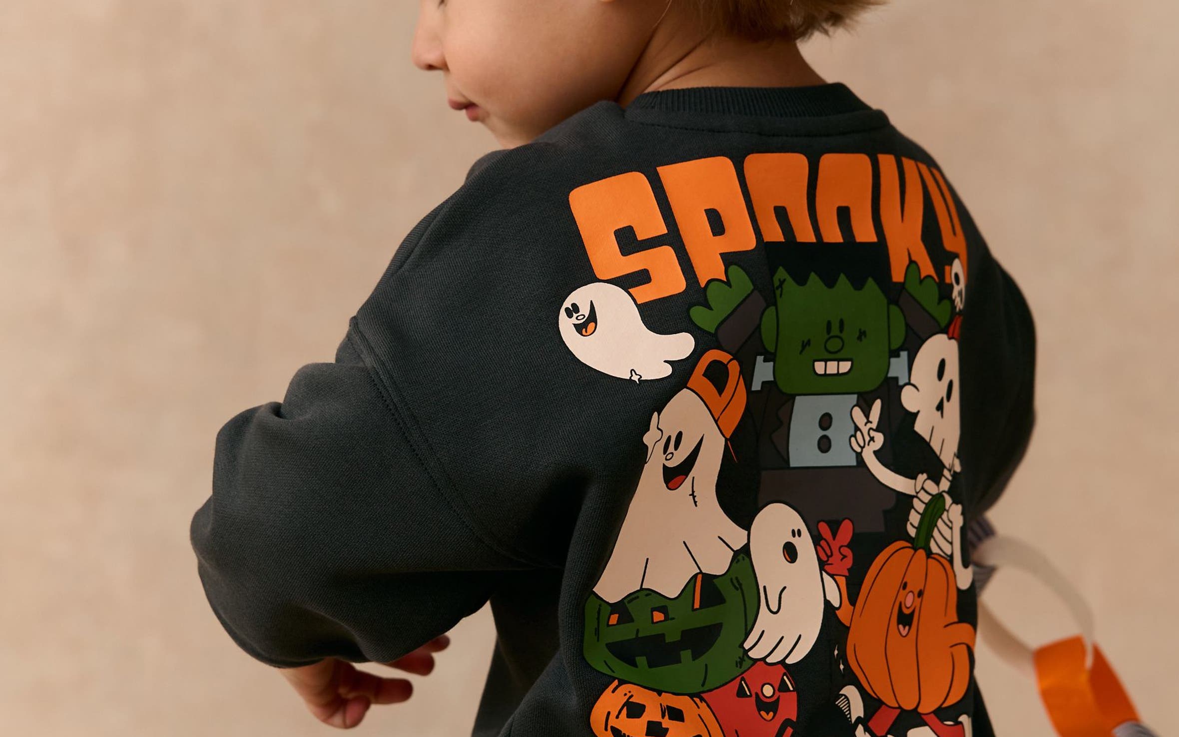 A boy in a black graphic sweatshirt with "Spooky Season" text and cartoon ghost, pumpkin and monster characters on the back. 