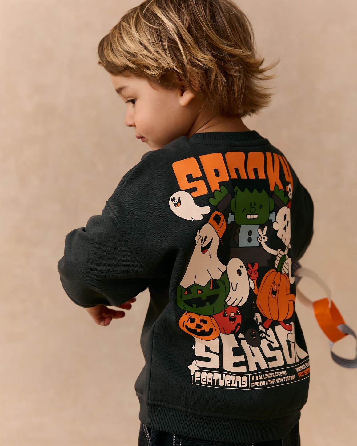 A boy in a black graphic sweatshirt with "Spooky Season" text and cartoon ghost, pumpkin and monster characters on the back. 