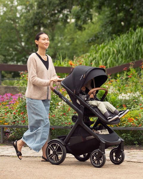 Woman walking a baby in a Nuna stroller past a flower garden.