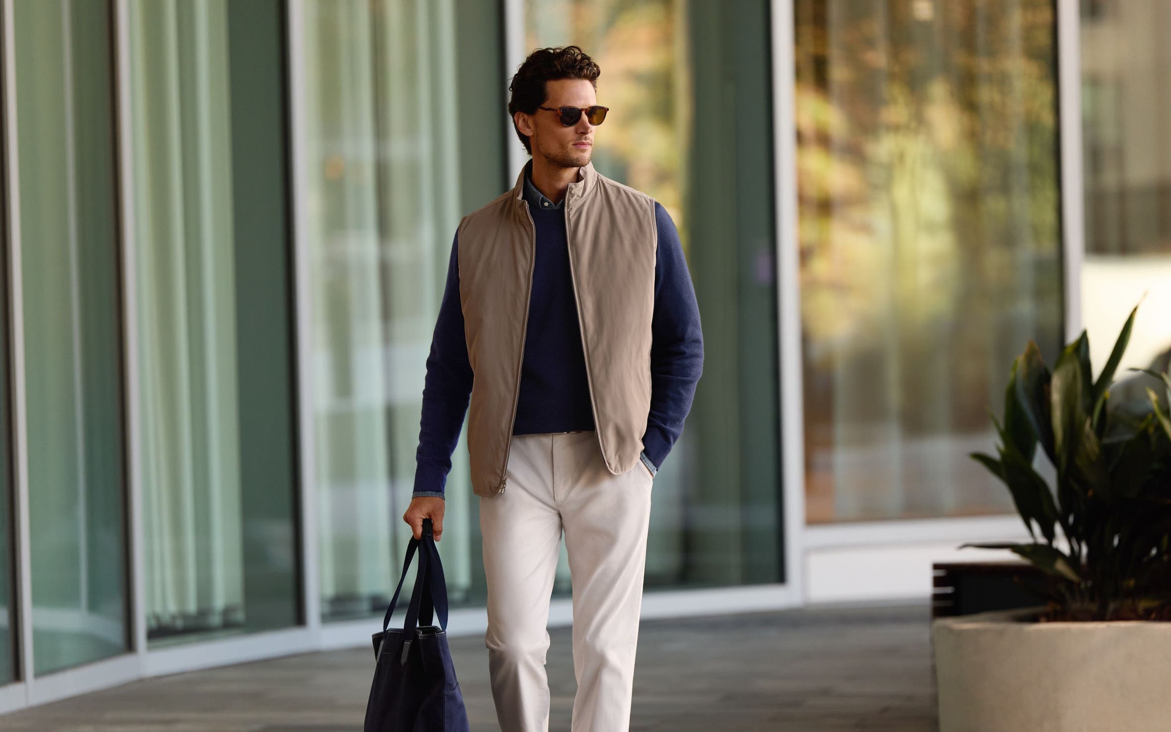 A man wearing a tan vest over a navy sweater with trousers.