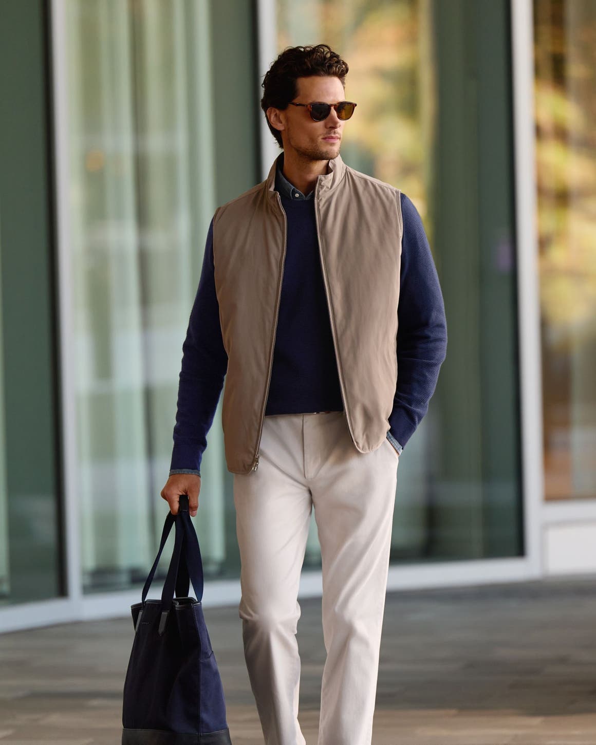 A man wearing a tan vest over a navy sweater with trousers.