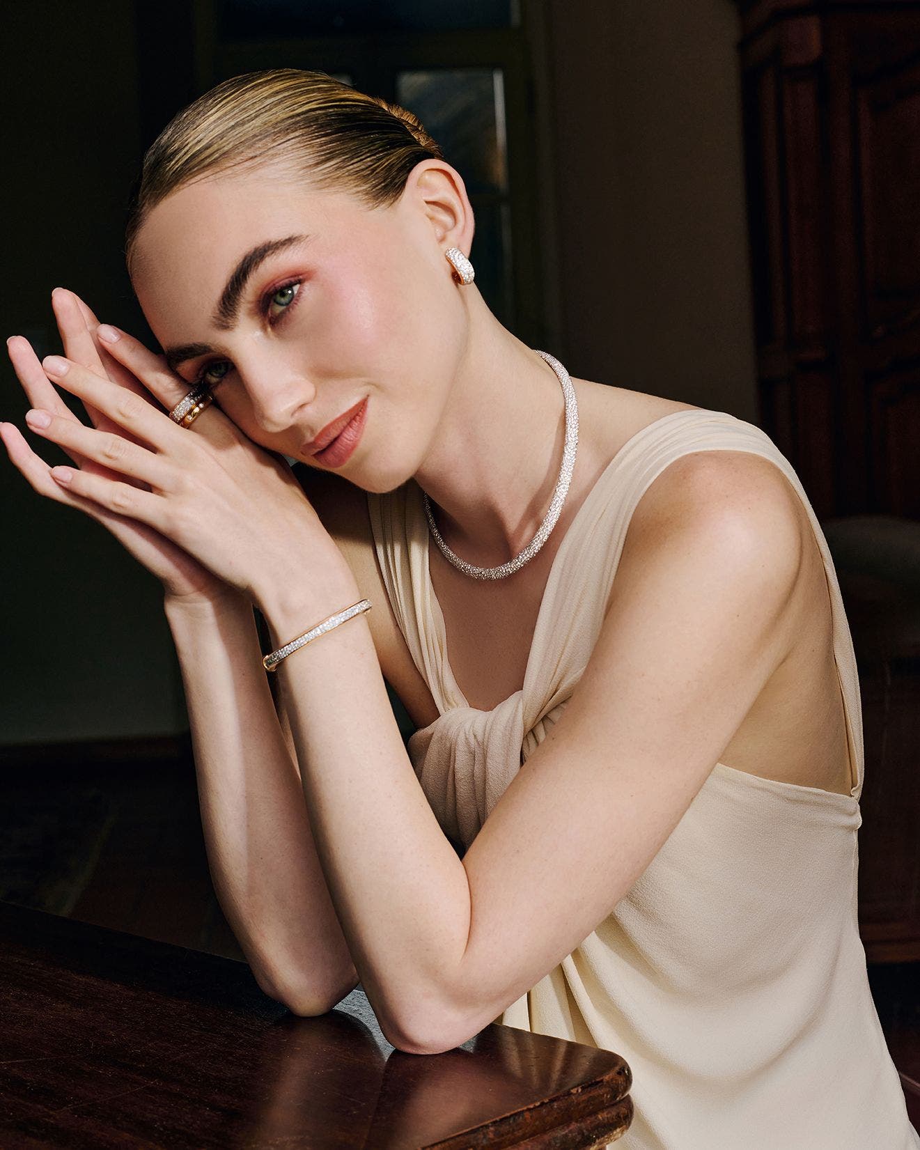 A woman sitting at a table wearing diamond earrings, a necklace, rings and a bracelet.