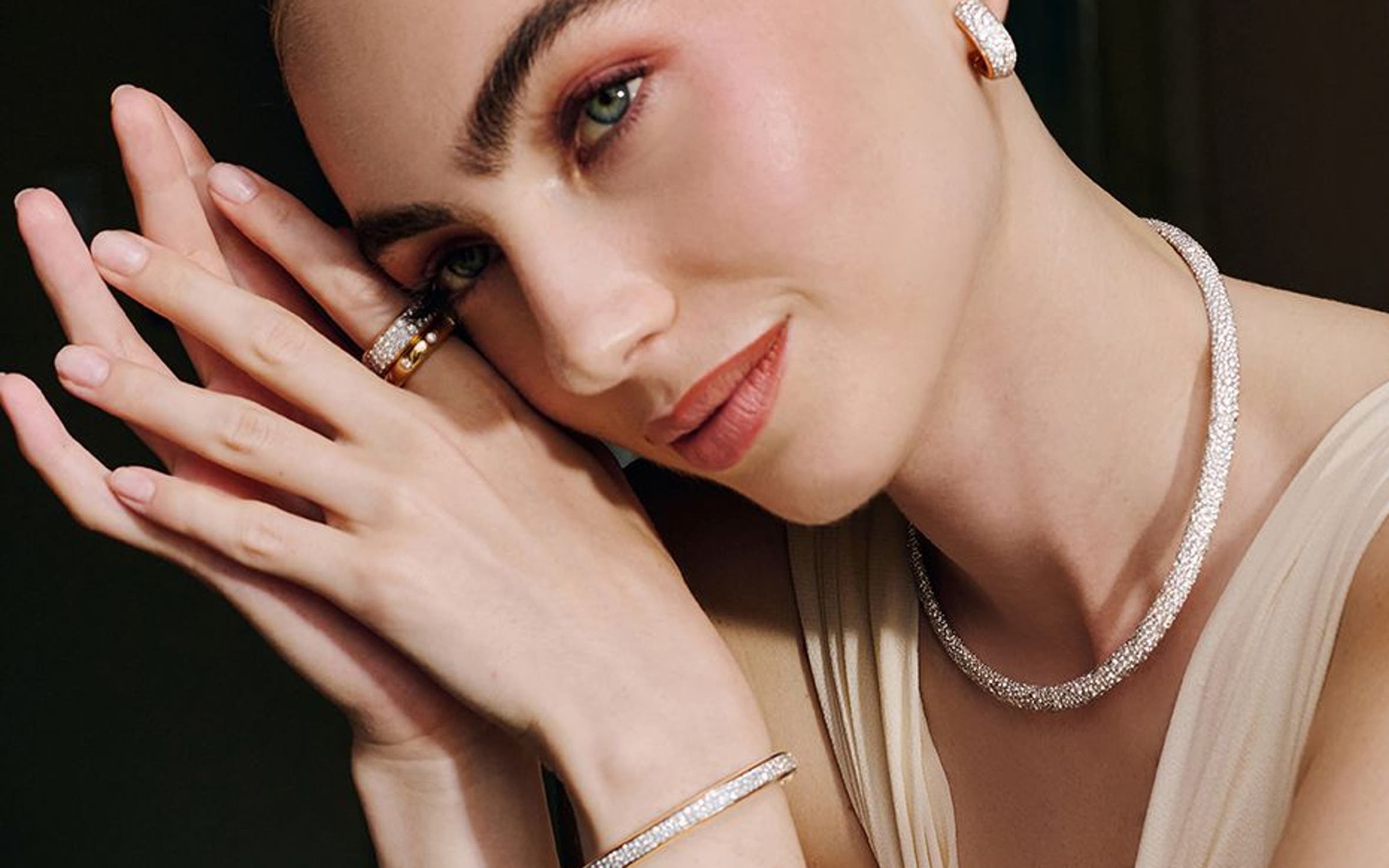 A woman sitting at a table wearing diamond earrings, a necklace, rings and a bracelet.