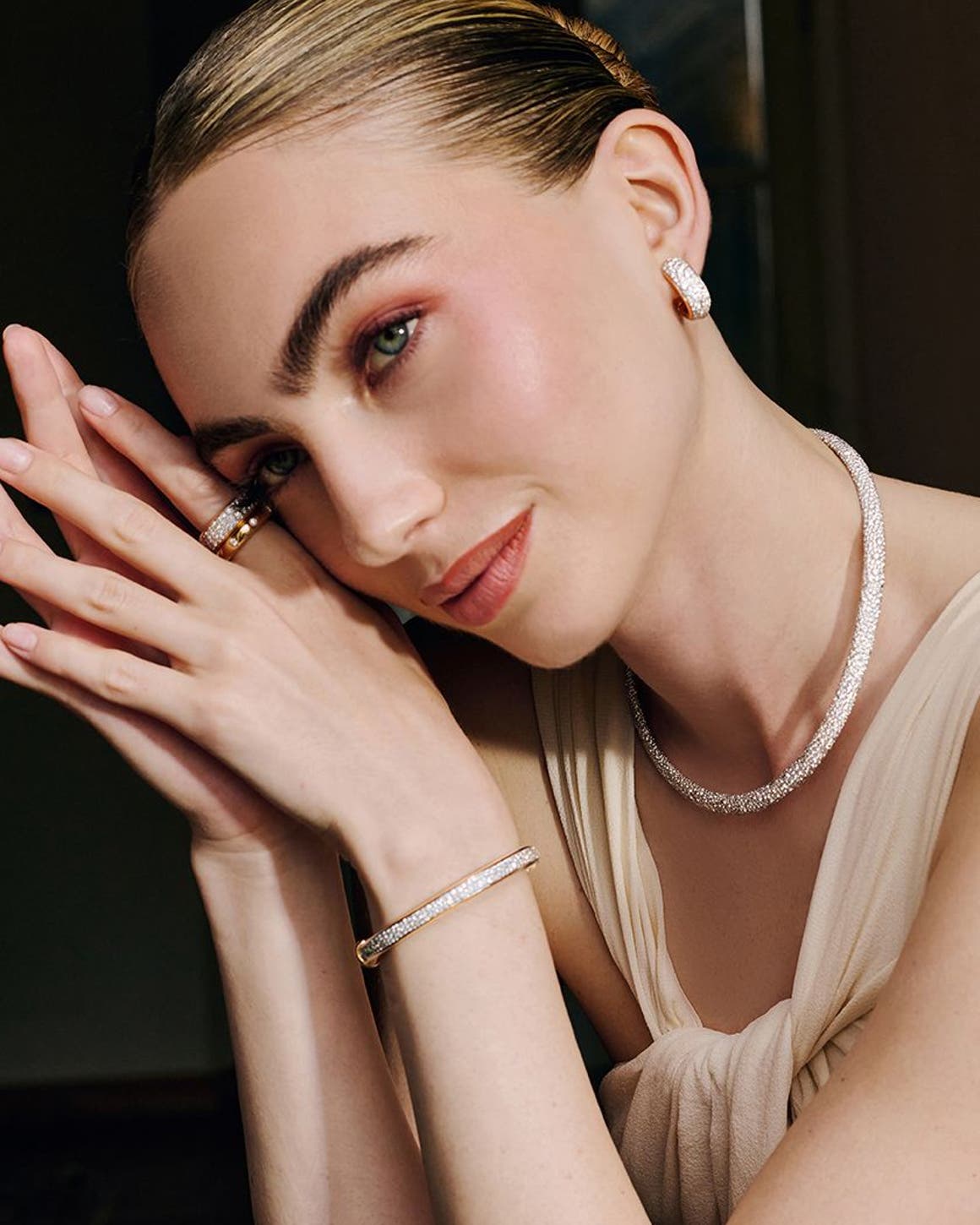 A woman sitting at a table wearing diamond earrings, a necklace, rings and a bracelet.