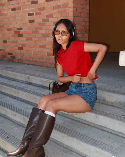 A woman wearing a red top, jean shorts, knee-high boots, sunglasses and headphones with a handbag.