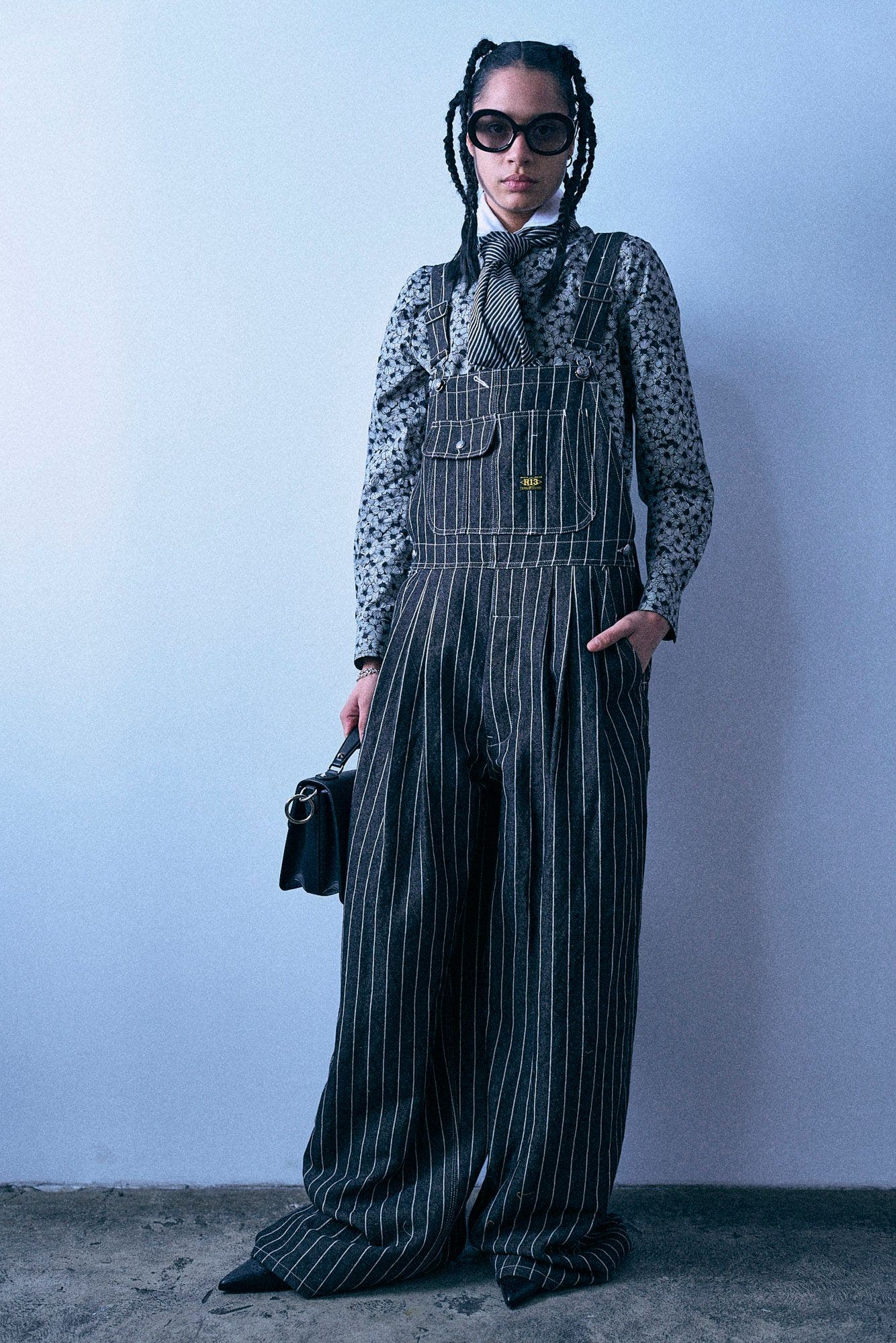 A woman with braided hair and black sunglasses stands against a plain light gray background, wearing a black-and-white polka-dot blouse under black-and-white pinstriped overalls, a black neck scarf, and holding a small black handbag.