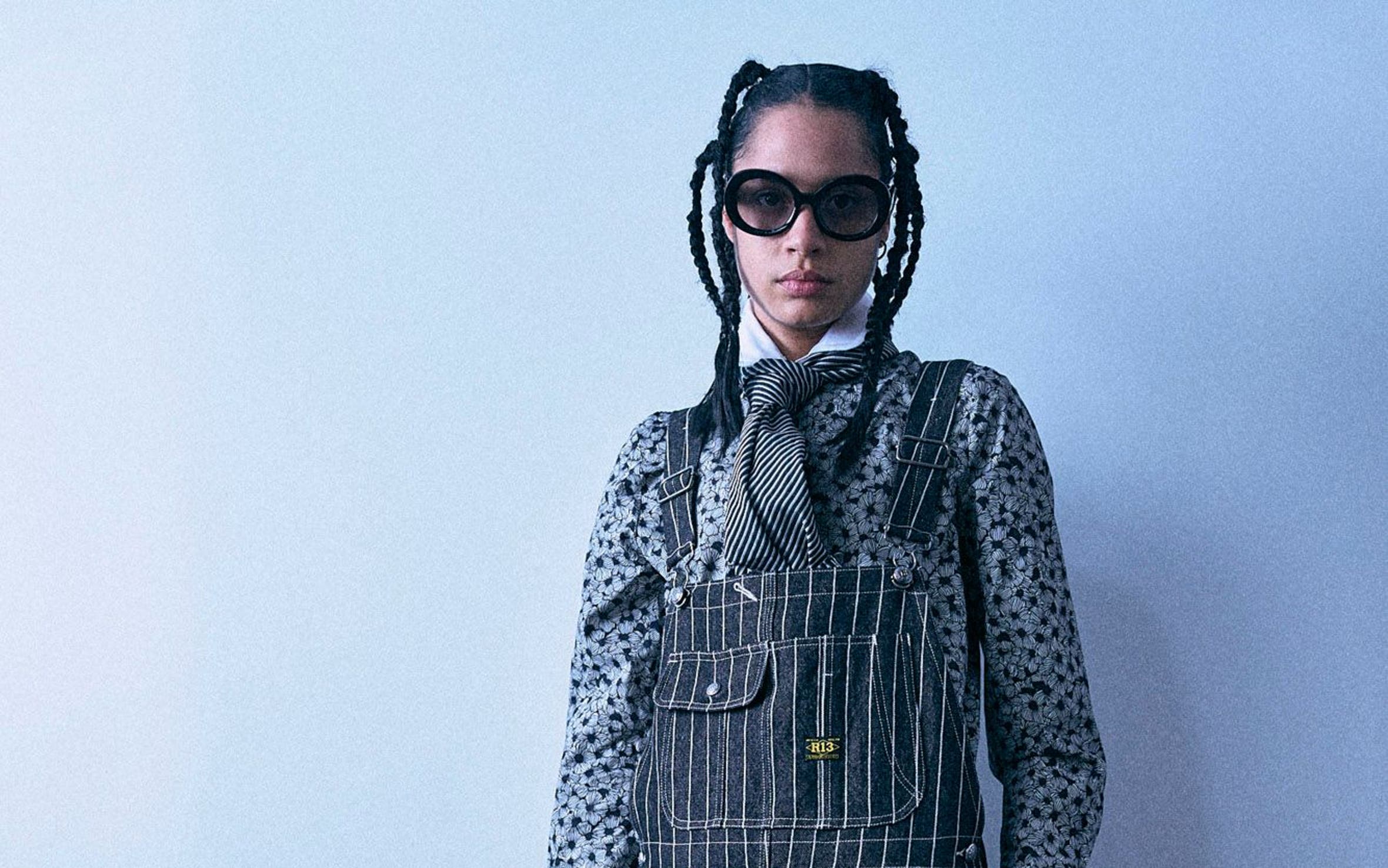 A woman with braided hair and black sunglasses stands against a plain light gray background, wearing a black-and-white polka-dot blouse under black-and-white pinstriped overalls, a black neck scarf, and holding a small black handbag.