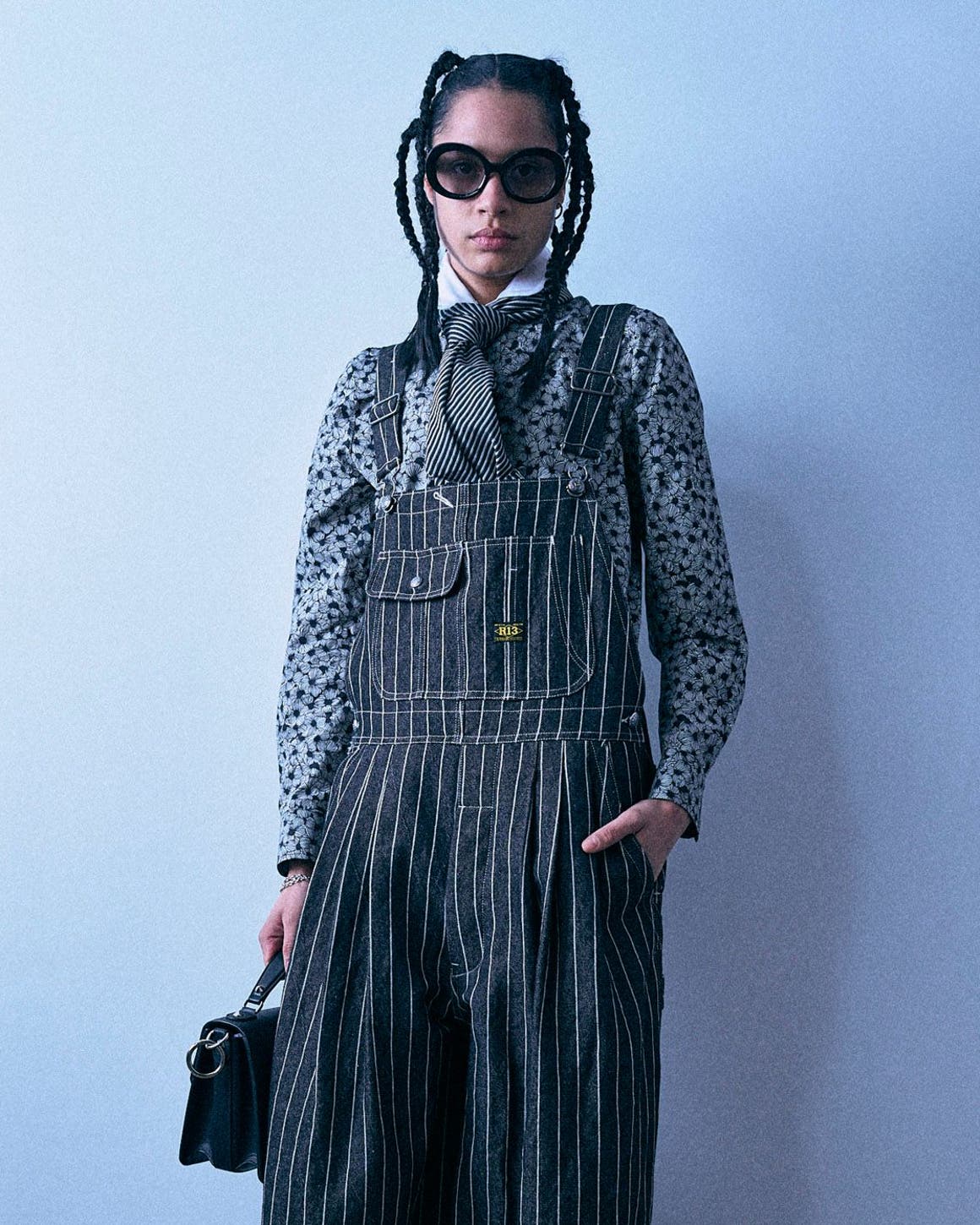 A woman with braided hair and black sunglasses stands against a plain light gray background, wearing a black-and-white polka-dot blouse under black-and-white pinstriped overalls, a black neck scarf, and holding a small black handbag.
