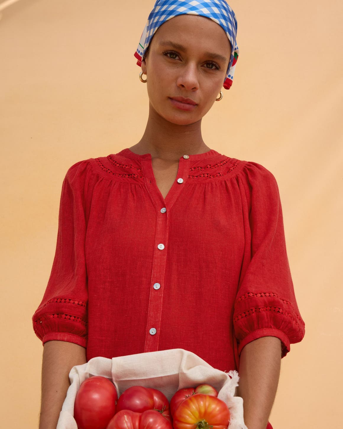 A woman wearing a gingham head scarf and red top carrying tomatoes.
